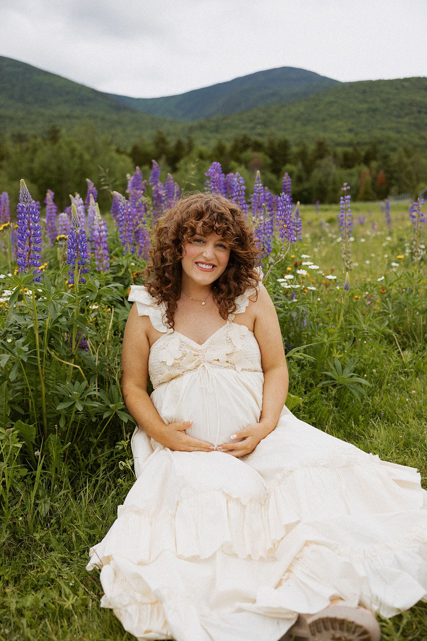 Pregnant woman in white dress sitting in lupine wildflower field in New Hampshire taking motherhood photos