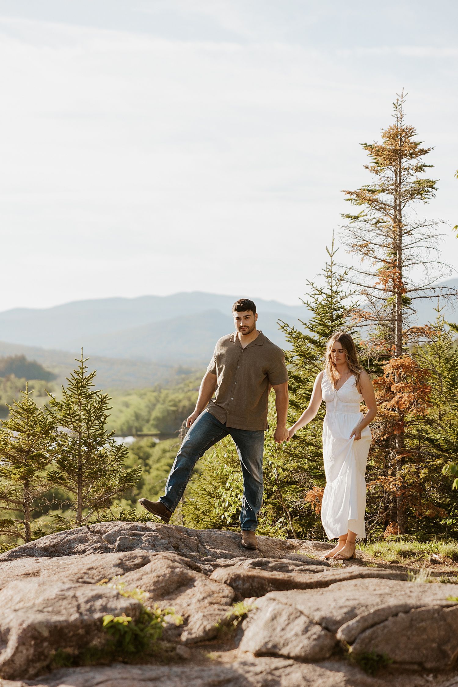 Couple holding hands on rocky mountain summit surrounded by pine trees with scenic valley and mountain views behind them.