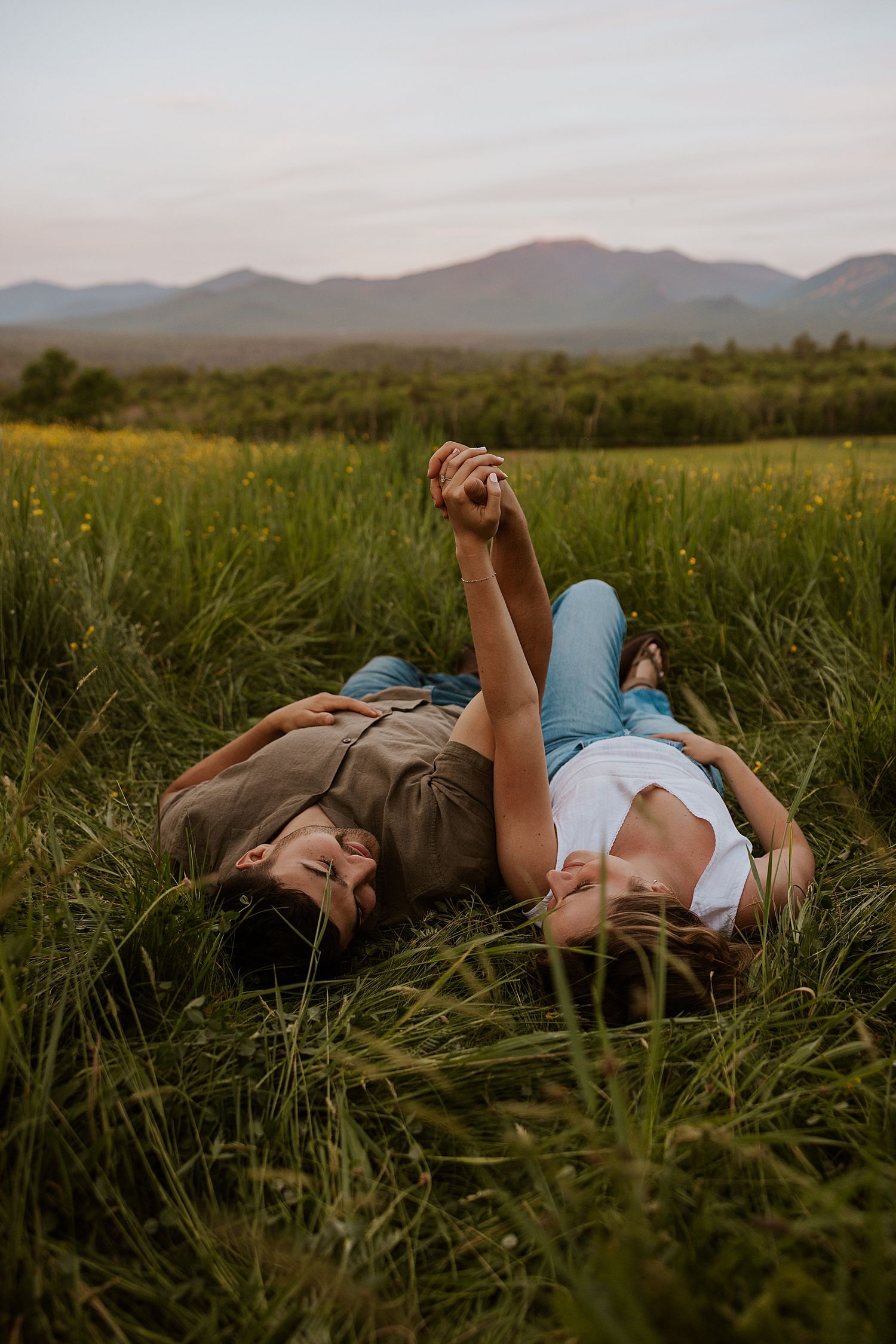 Couple lying in tall green grass holding hands with mountain landscape in background at dusk.