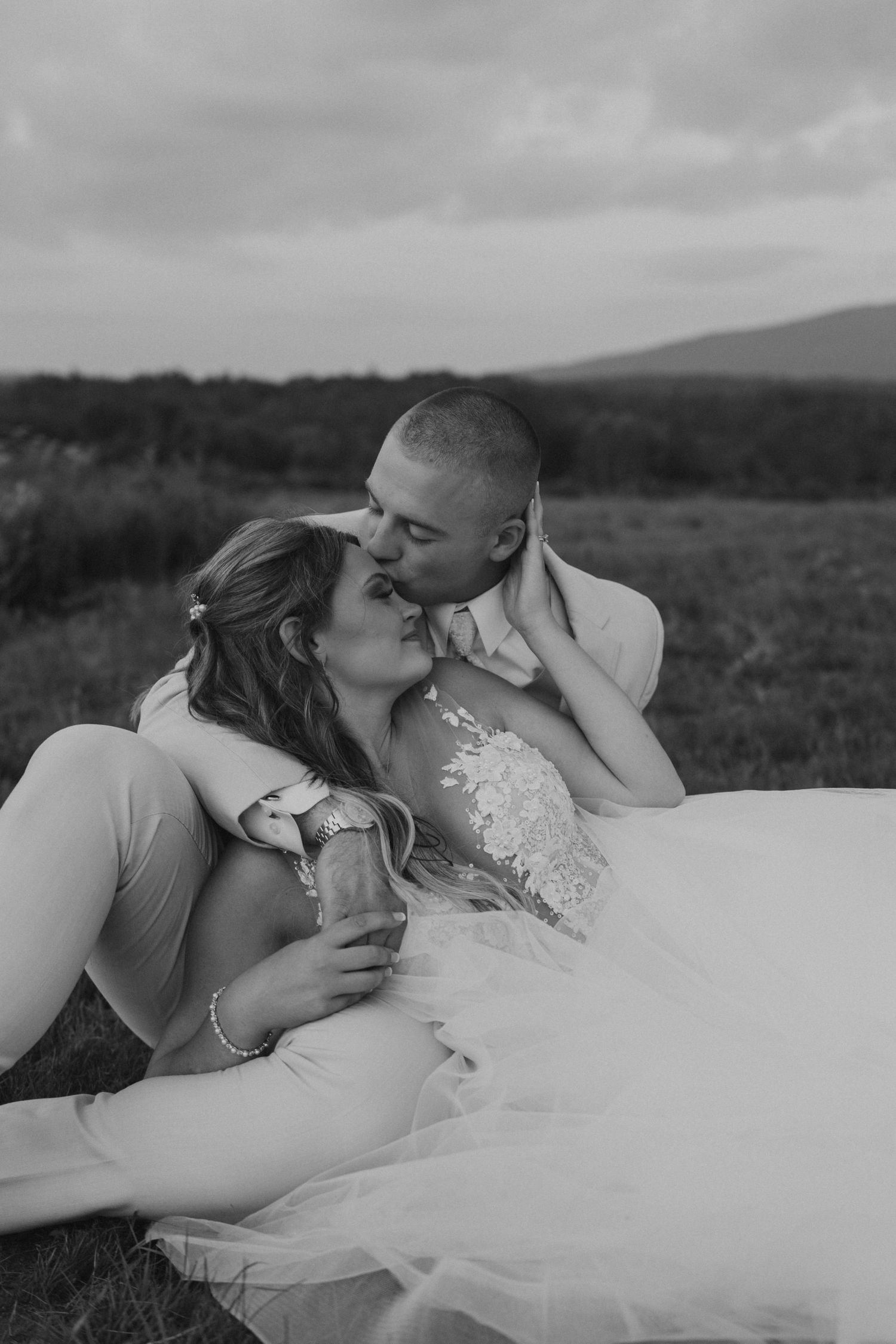 Black and white photo of bride and groom kissing while seated in a grassy field with mountains behind.