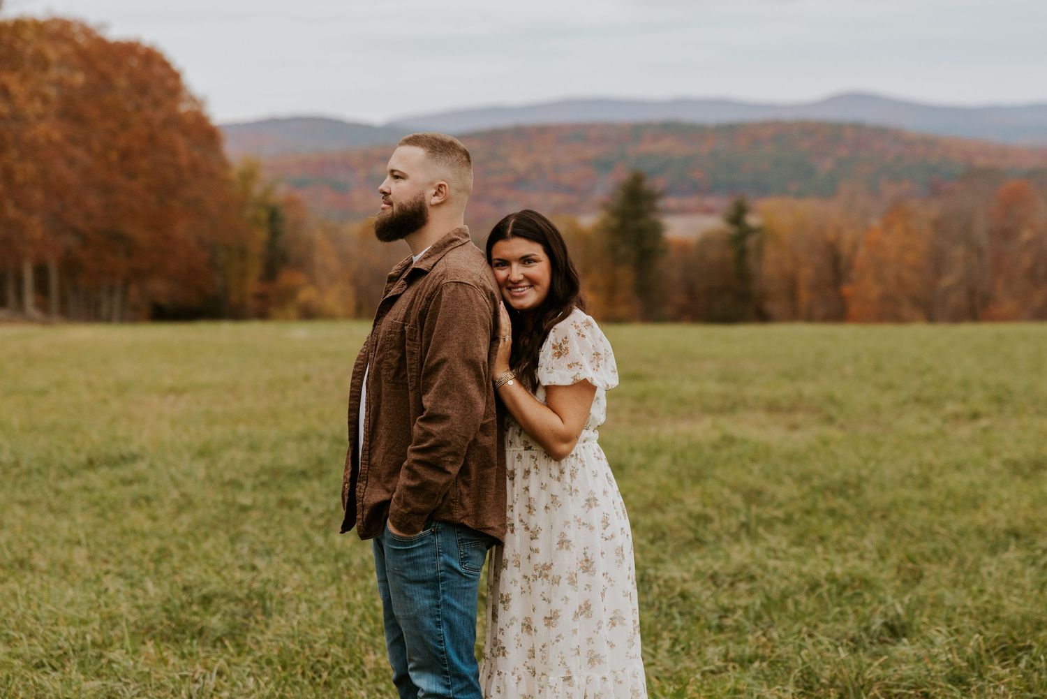 Couple posing back-to-back in a fall field, woman in floral dress smiling at camera with mountain backdrop.