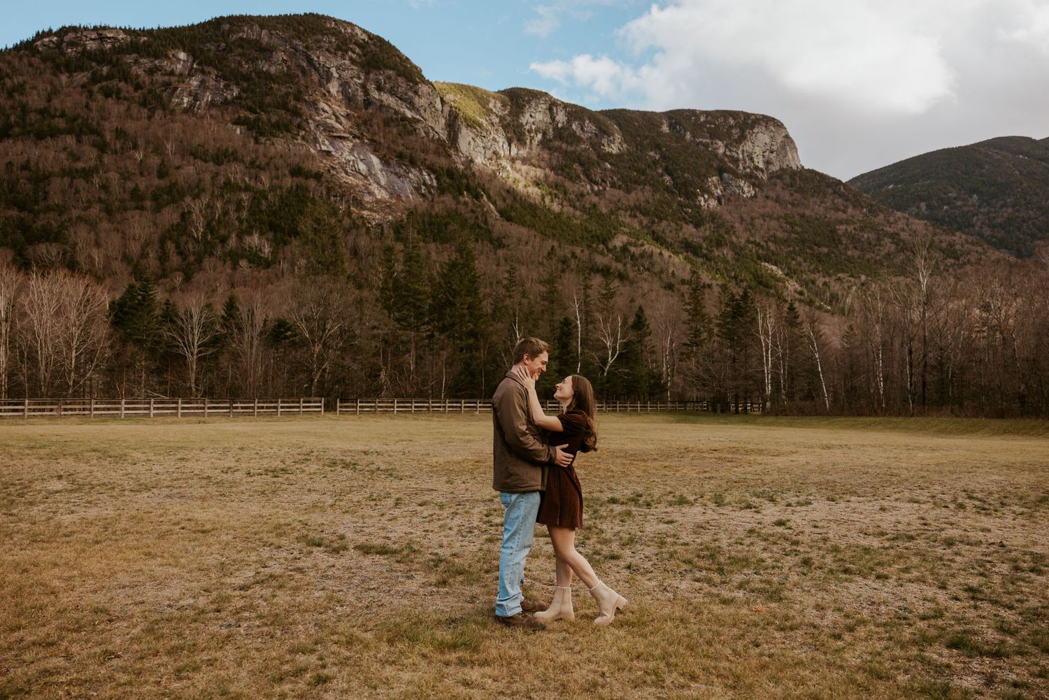 Couple embracing and dancing in an open mountain meadow with rocky cliffs and bare trees in early spring.