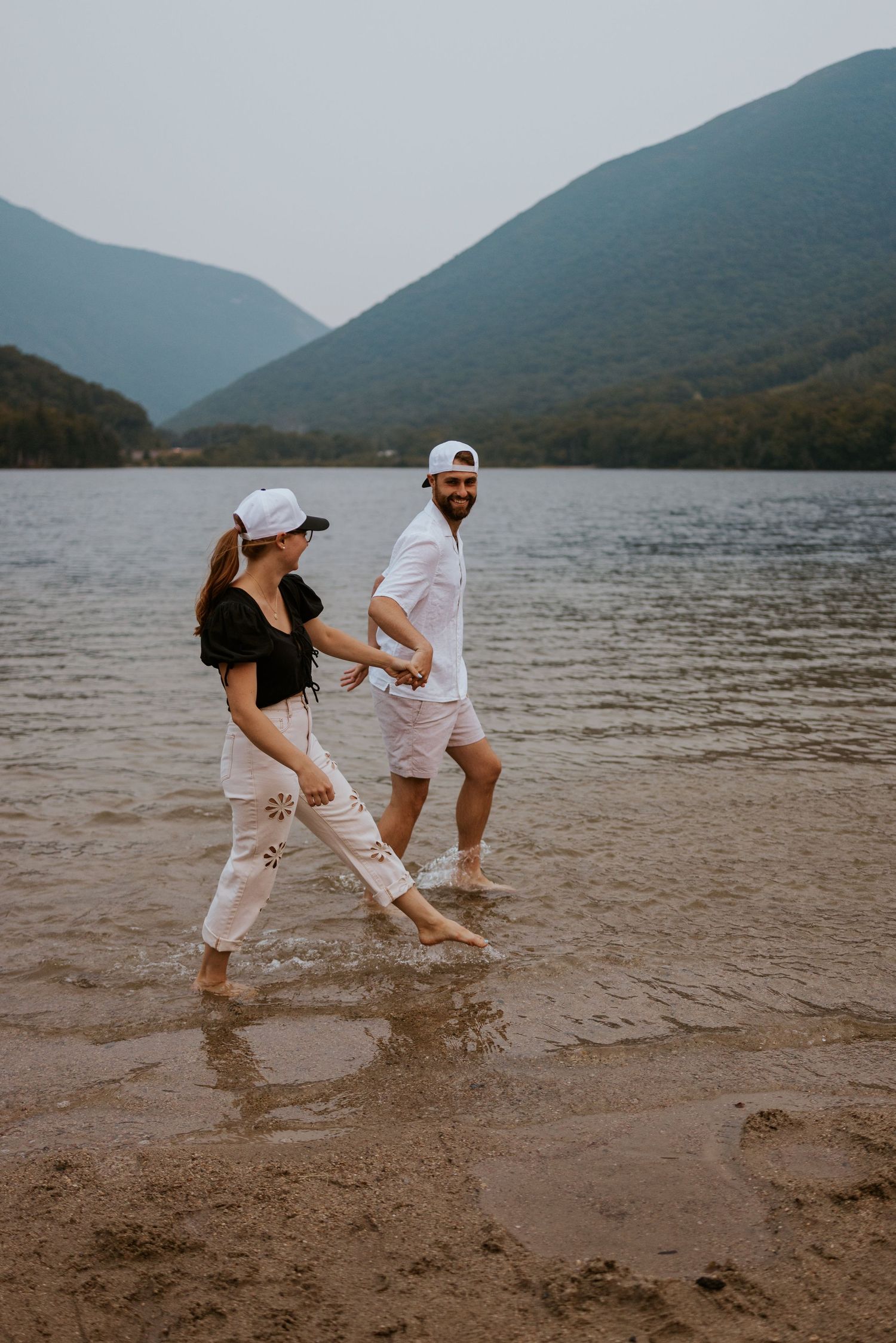 Couple walking hand in hand through shallow lake water with mountain scenery behind them.