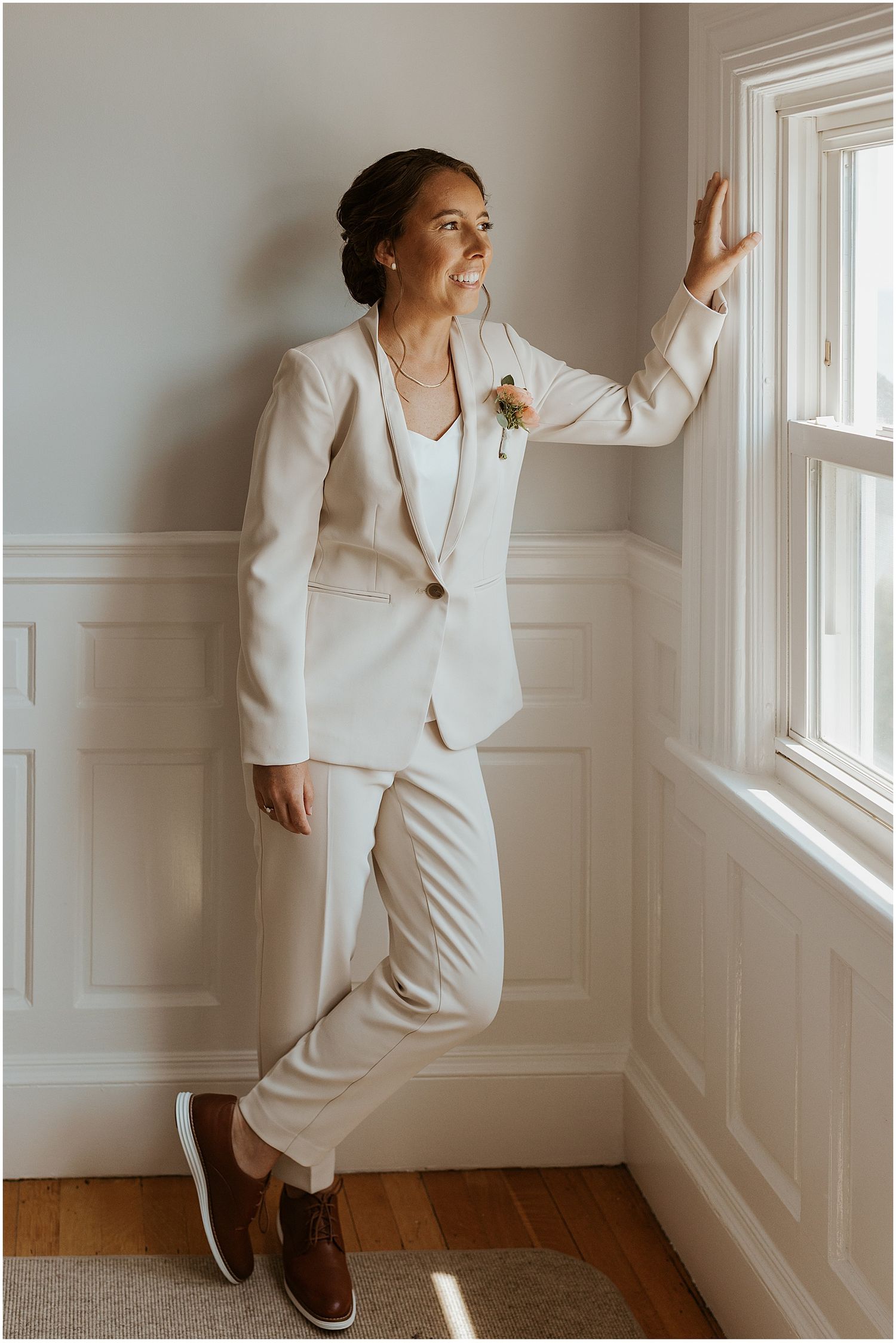 A figure in a crisp white pantsuit and brown shoes poses playfully against a white wall near a window.