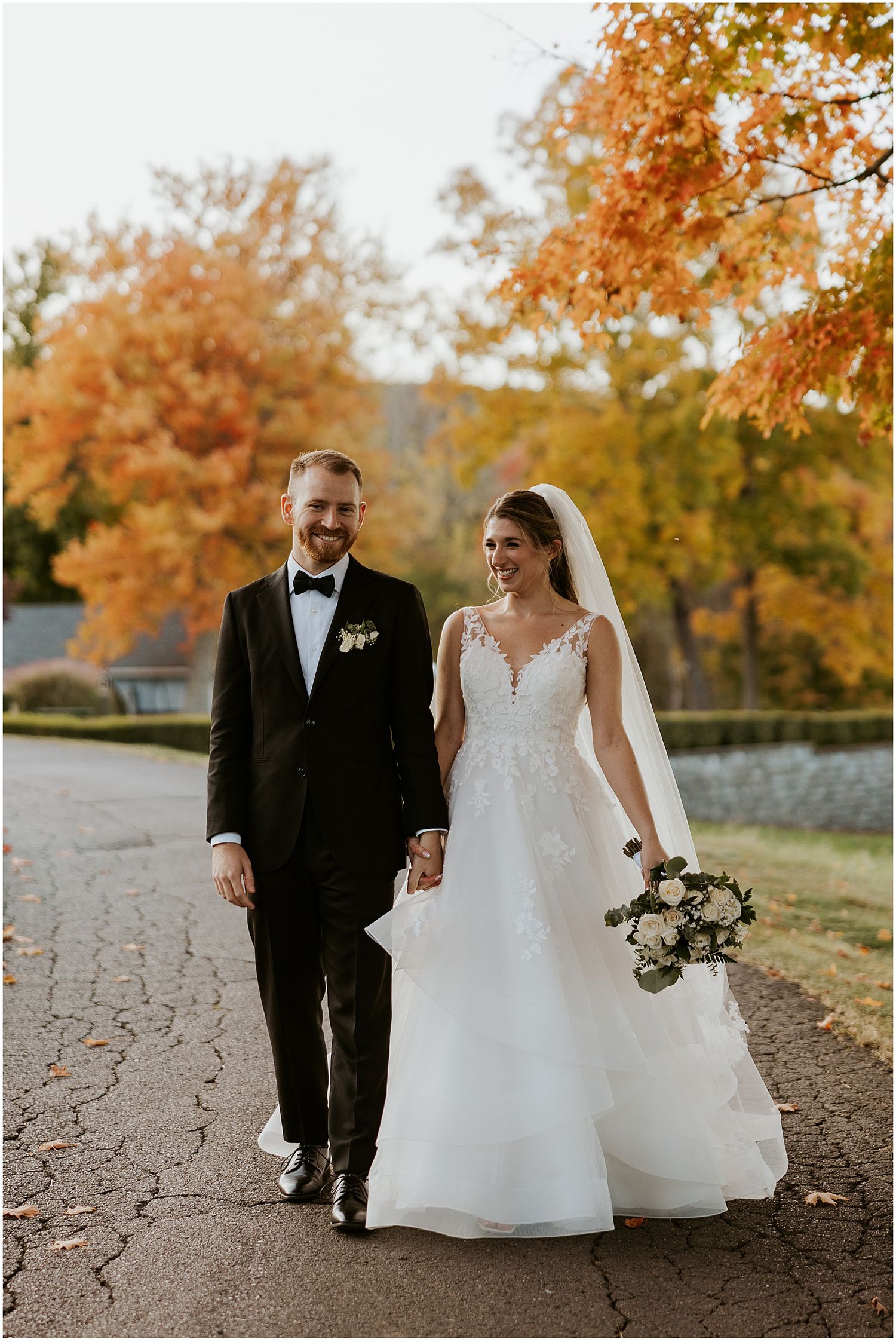 Bride and groom walking hand in hand on a path surrounded by beautiful fall foliage.