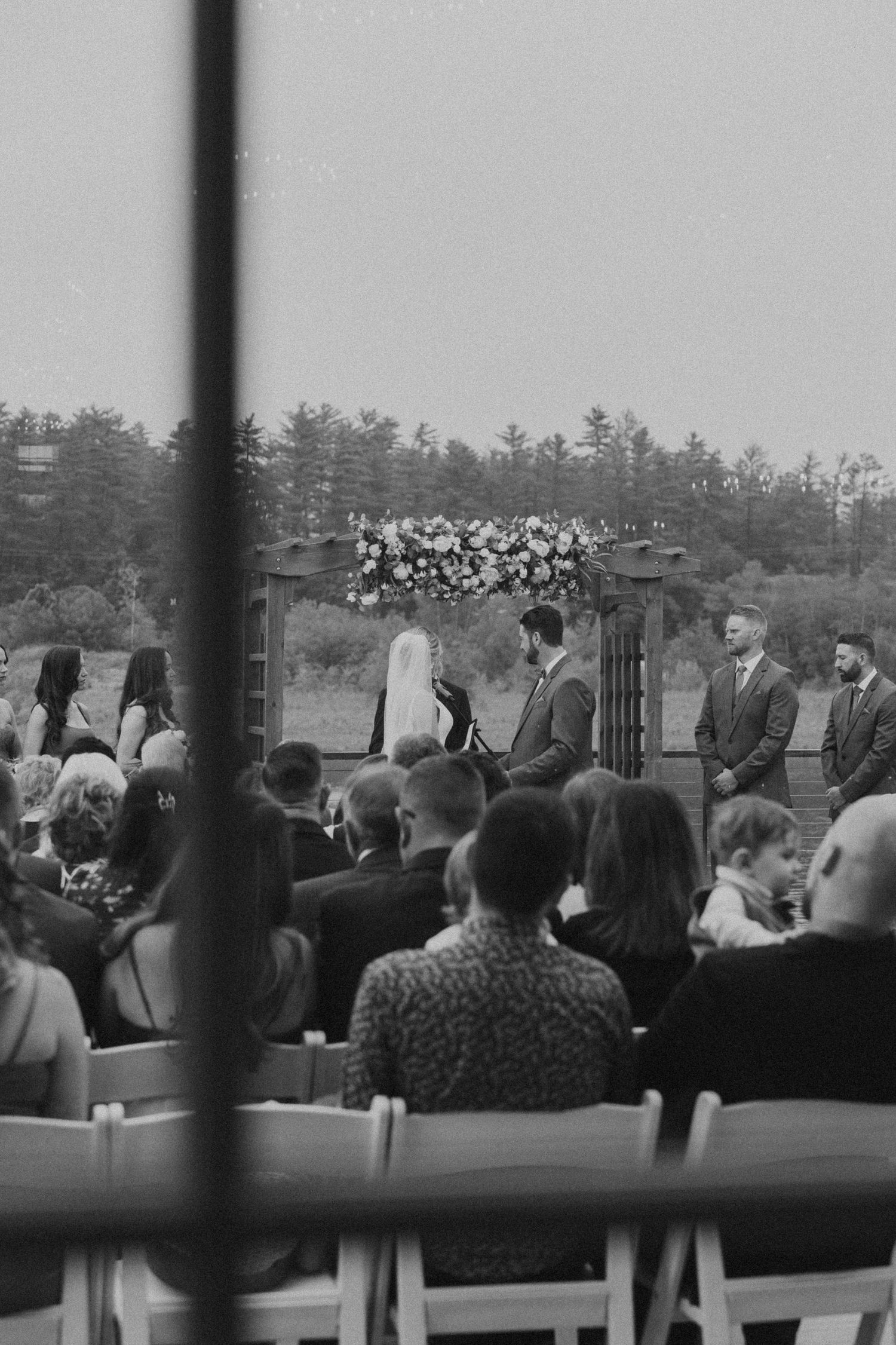 Black and white photo of outdoor wedding ceremony viewed through window with floral arch backdrop.