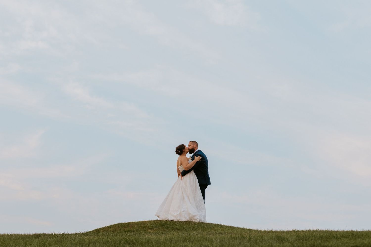 Bride and groom sharing a romantic kiss on a grassy hill under a soft blue sky on their wedding day.
