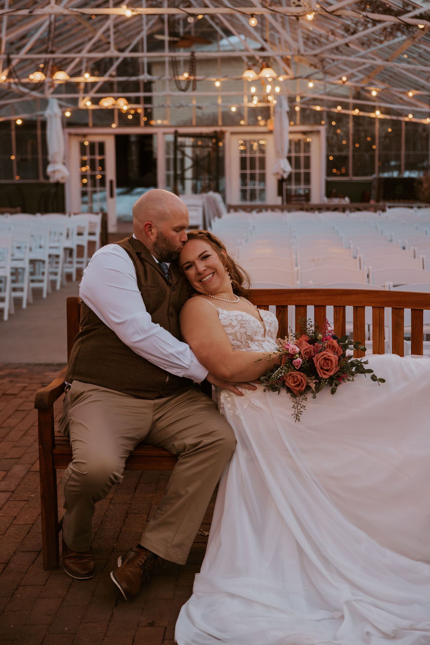 Newlyweds sharing a romantic moment on a wooden bench under string lights at dusk.
