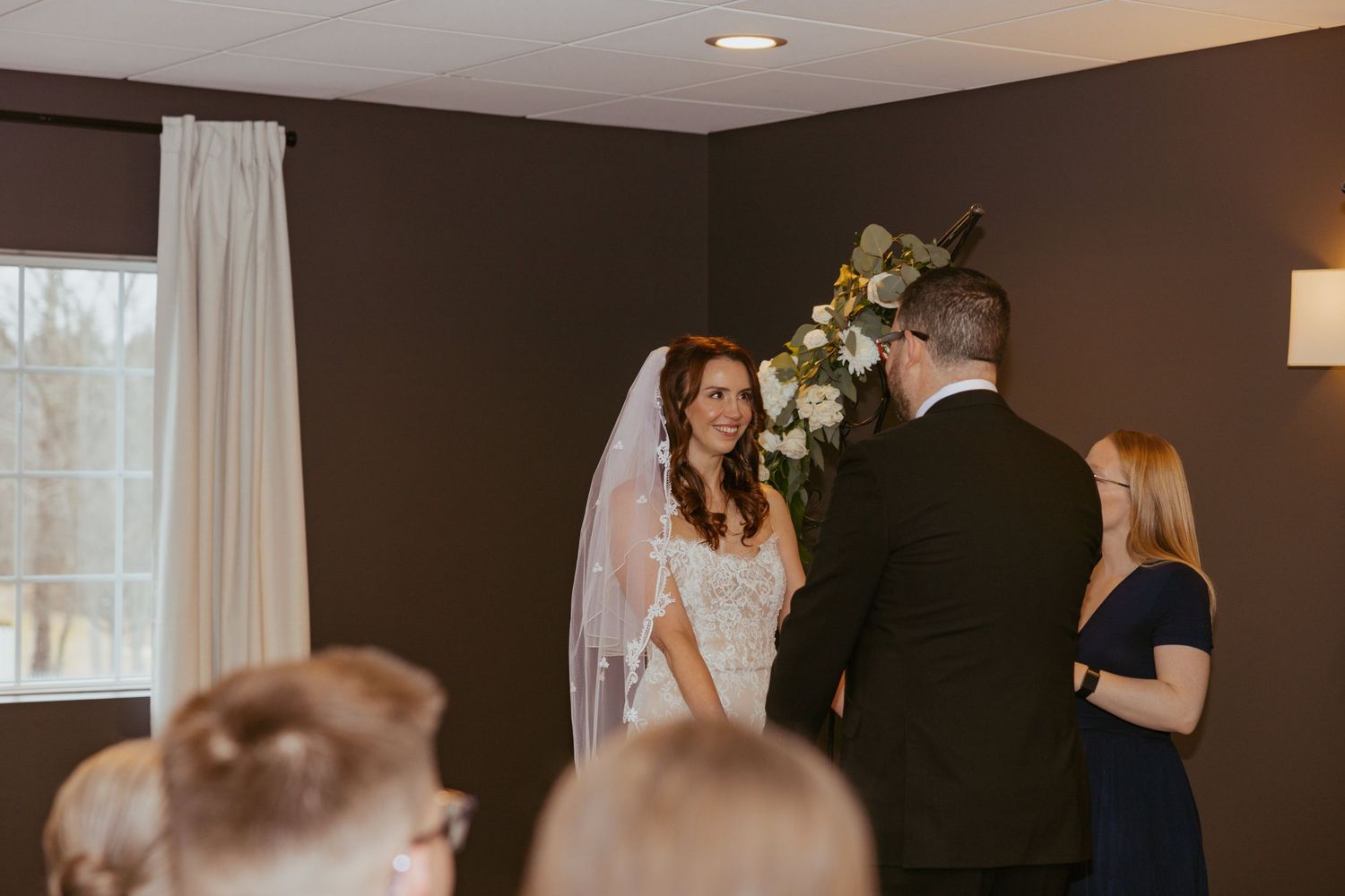Indoor wedding ceremony with a bride in white dress and veil standing with officiant and guests in attendance.