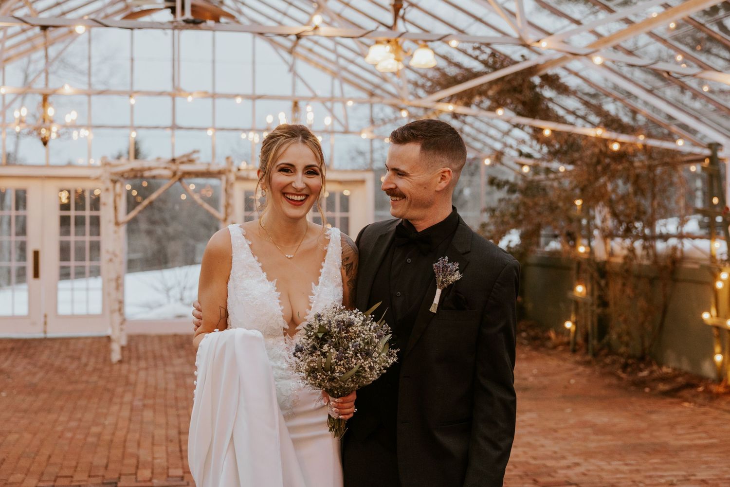 Smiling bride and groom in black suit posing inside warmly lit winter greenhouse venue.