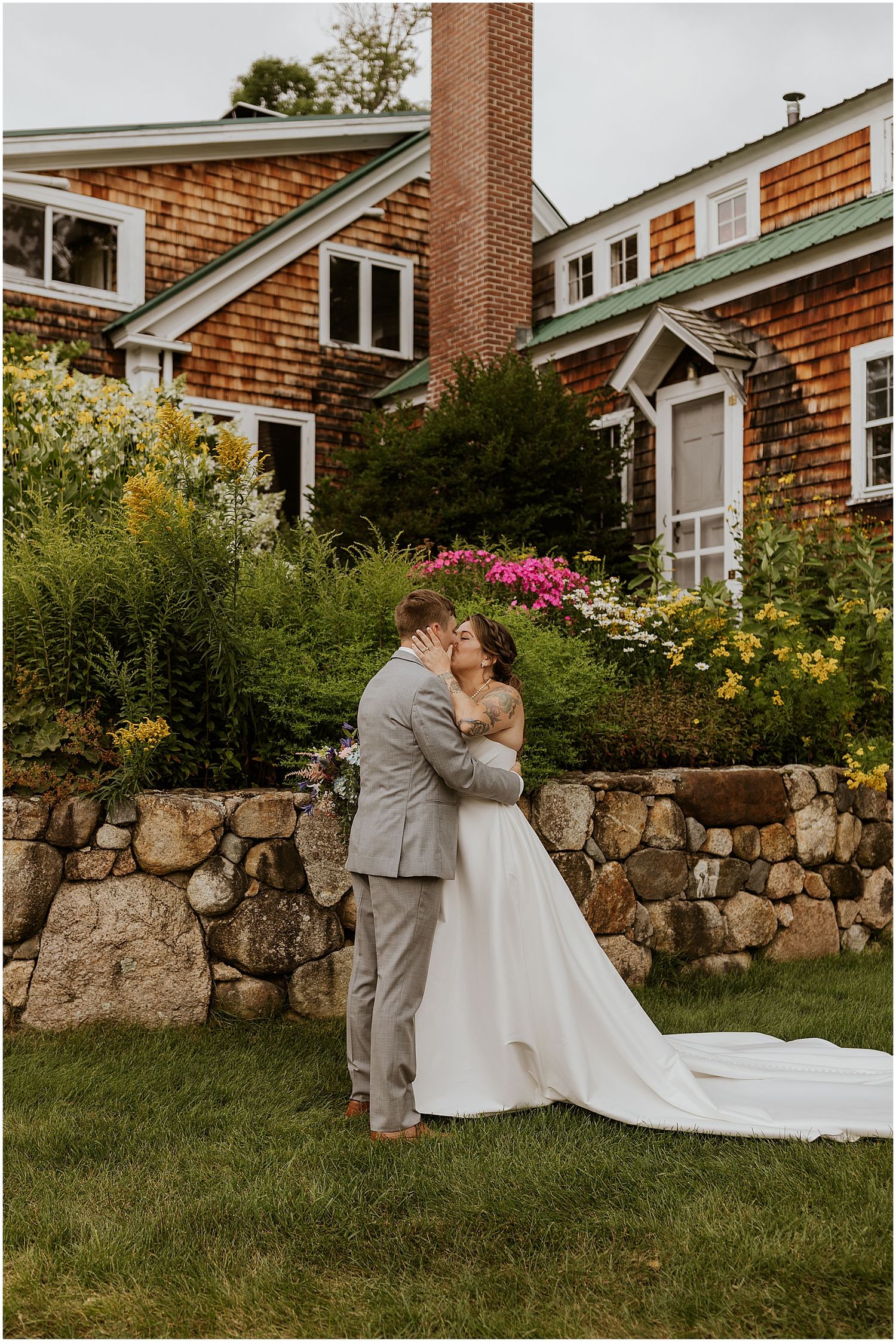 A romantic wedding moment in front of a rustic cedar-shingled house with colorful garden flowers and stone wall.
