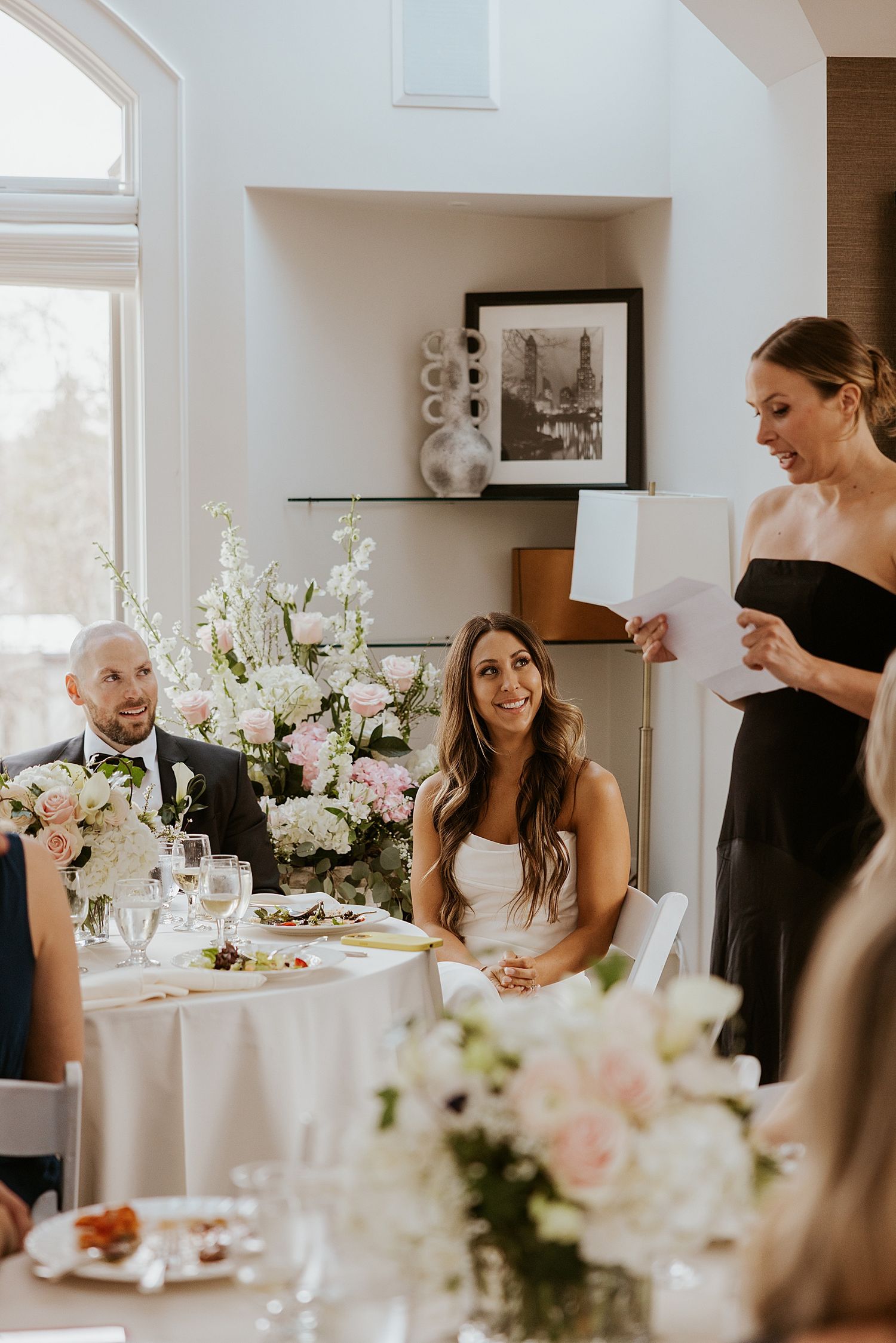 Bride in white dress smiles as bridesmaid in black gives a heartfelt wedding speech at an elegant floral reception.