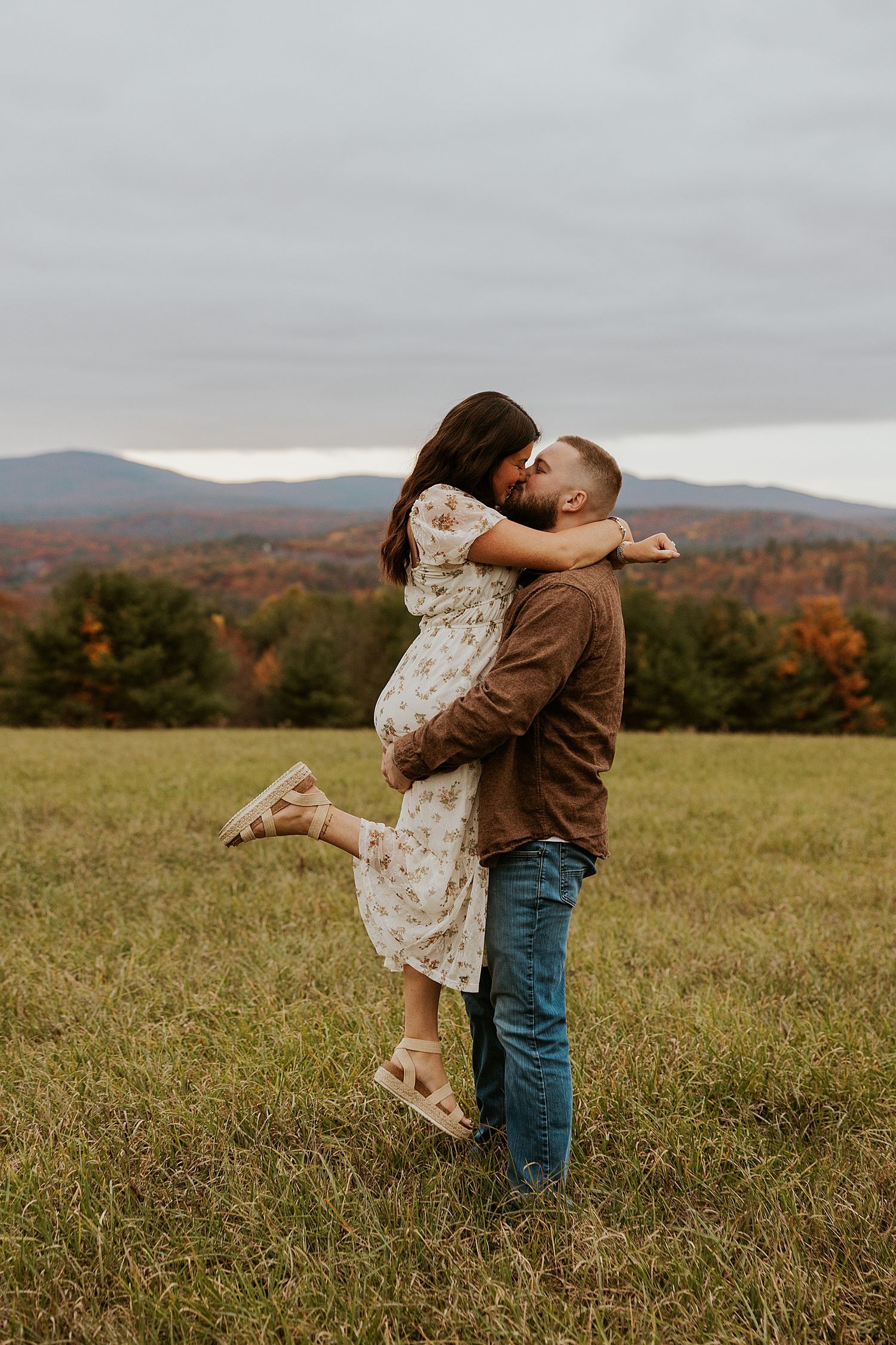 Couple sharing a romantic kiss in an autumn field, with colorful fall foliage and mountains in the background.