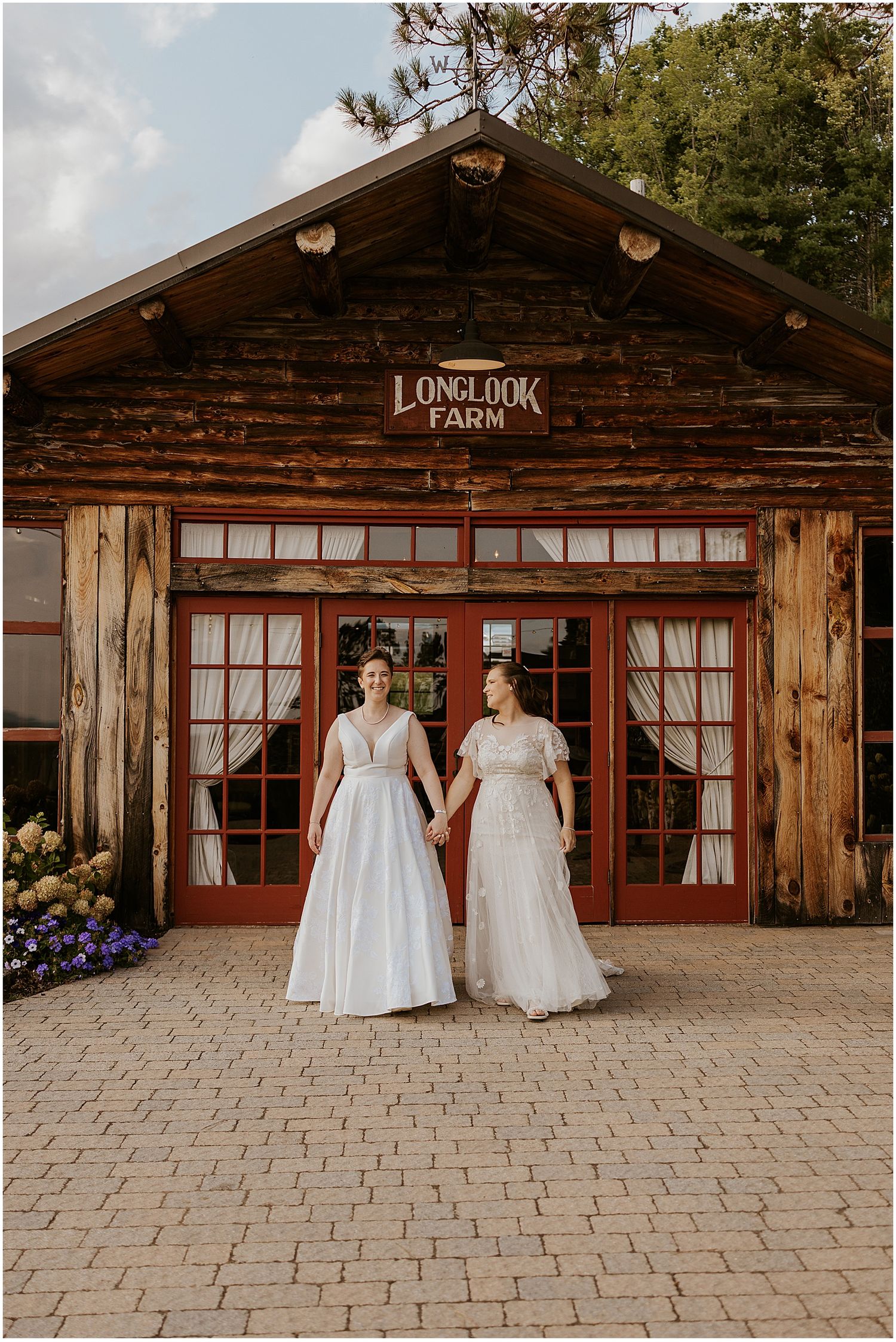 Two brides in white wedding dresses stand together outside a rustic wooden lodge venue with windows and stone patio.