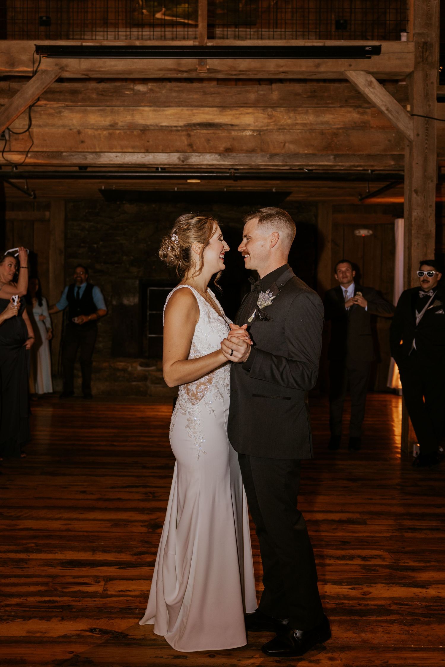 Bride and groom share a romantic first dance, smiling at each other in a warmly lit rustic barn venue.