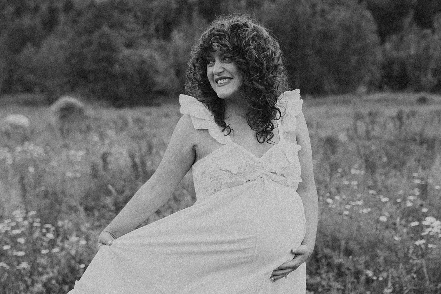 Smiling pregnant woman in white dress with curly hair posing in a field, black and white maternity photo.