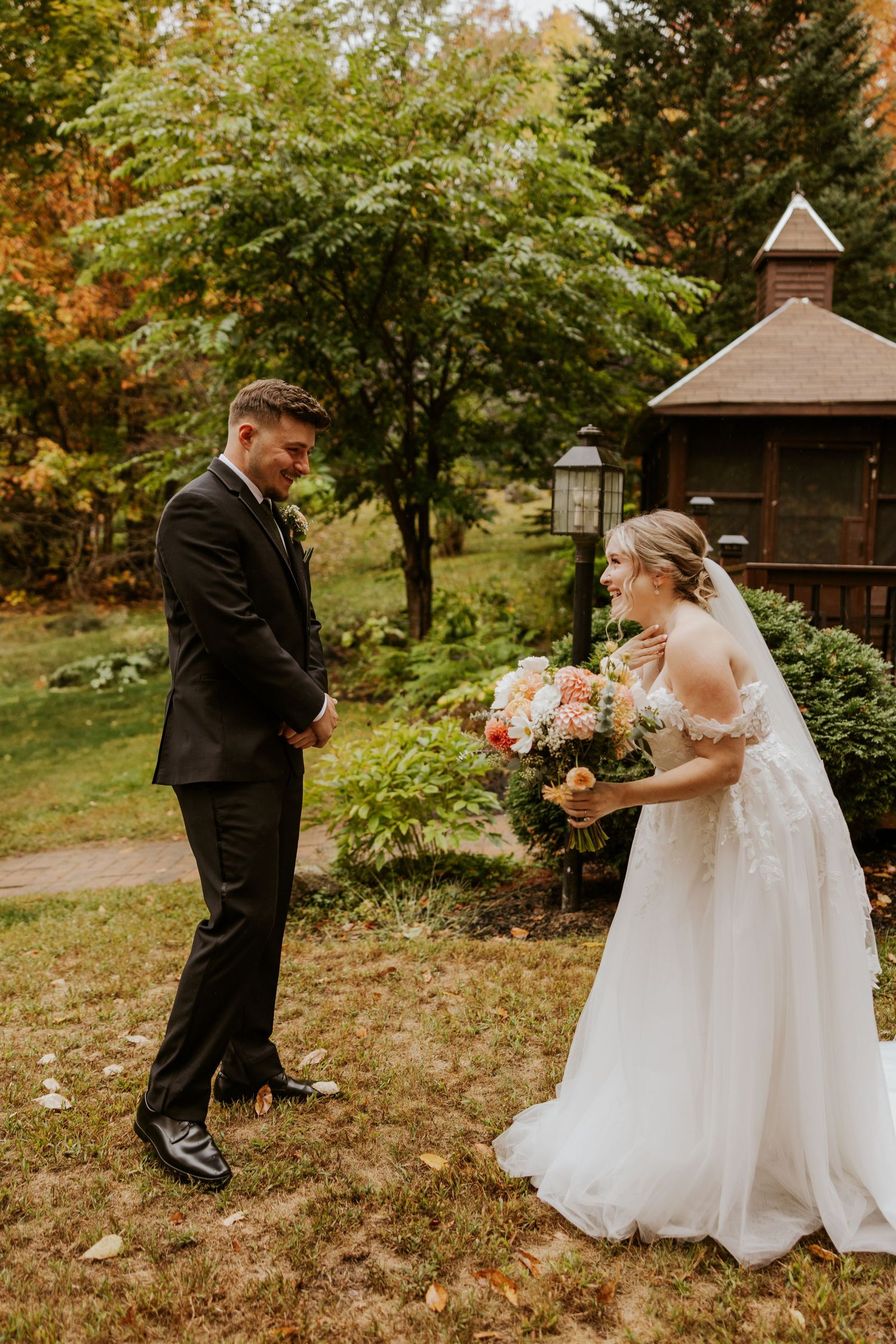 Bride and groom share joyful first look reaction outdoors near rustic gazebo with fall foliage backdrop.