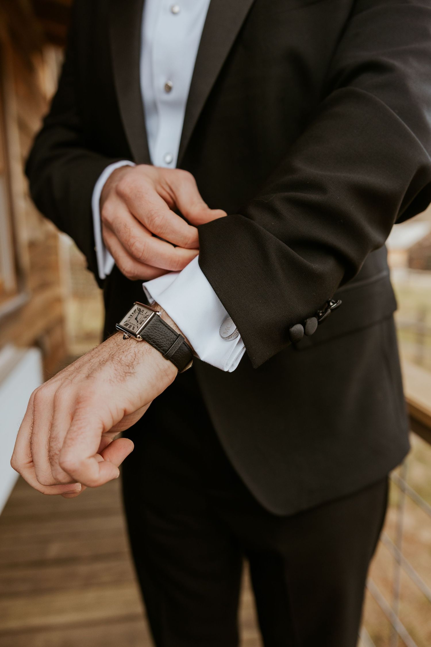 Groom in black tuxedo adjusting his cufflinks while wearing an elegant square-face leather watch.