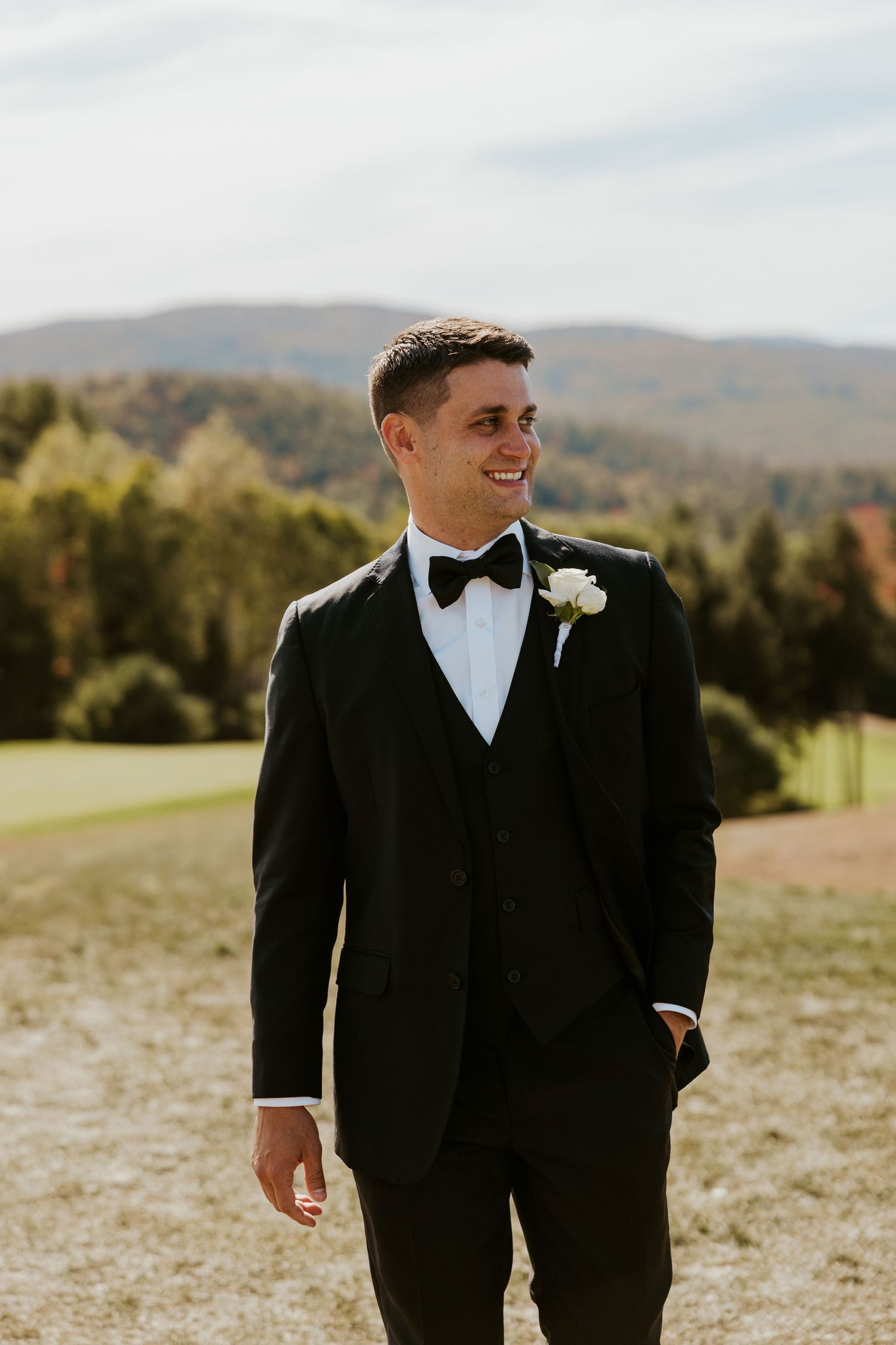 Groom in black tuxedo with white boutonniere smiling outdoors with mountain scenery behind him.