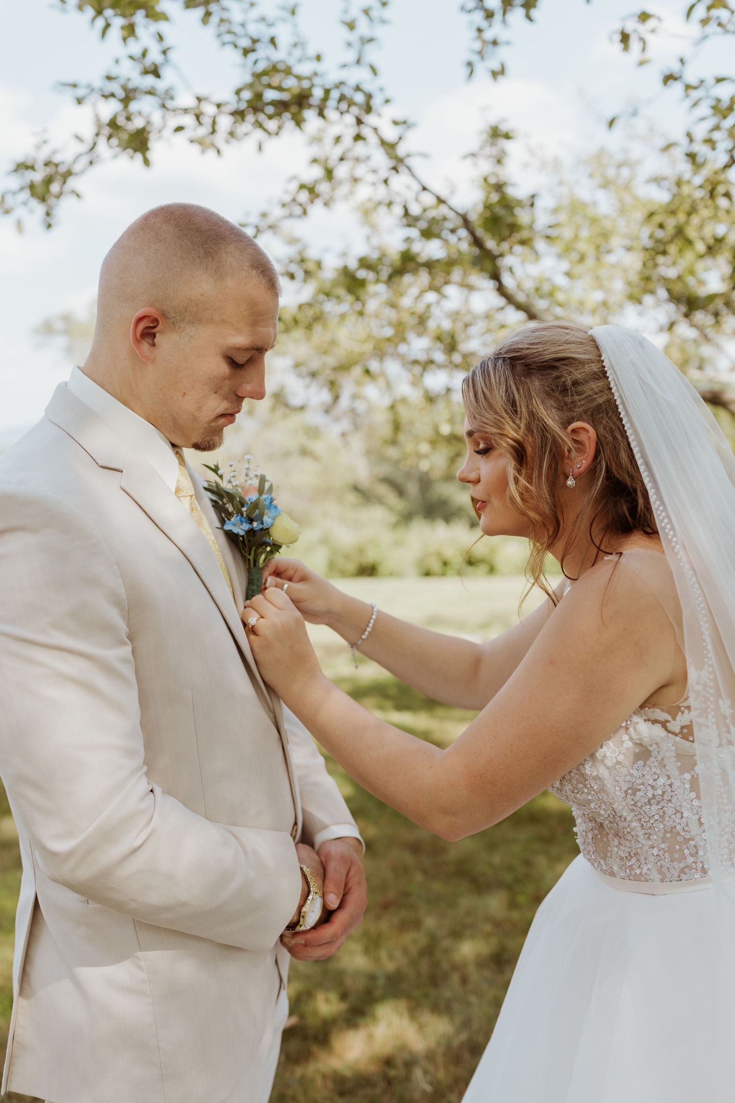 Bride carefully pins a floral boutonniere onto grooms beige suit jacket outdoors on wedding day.