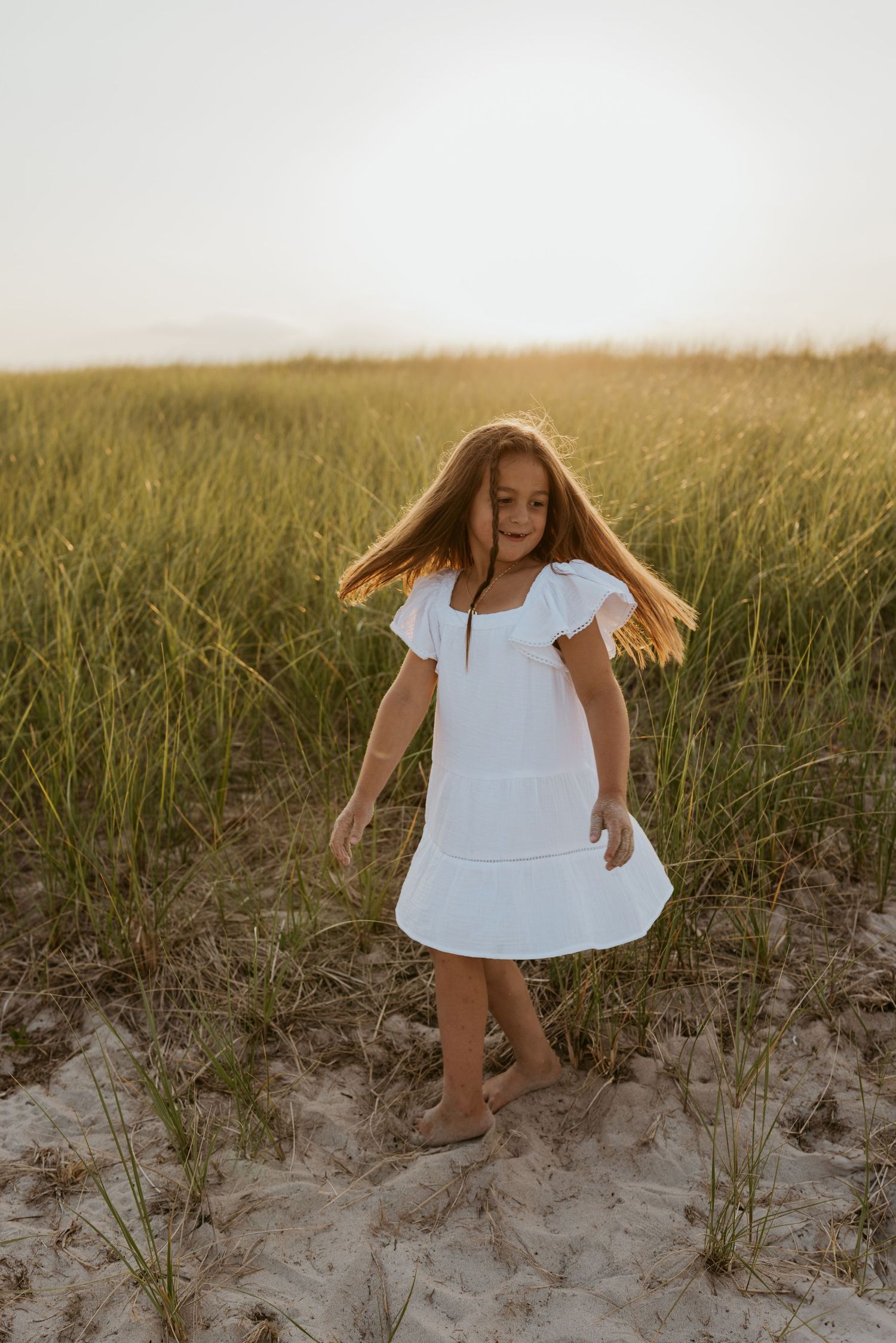 Young girl in white dress twirling on sandy beach with tall green grass at golden hour.