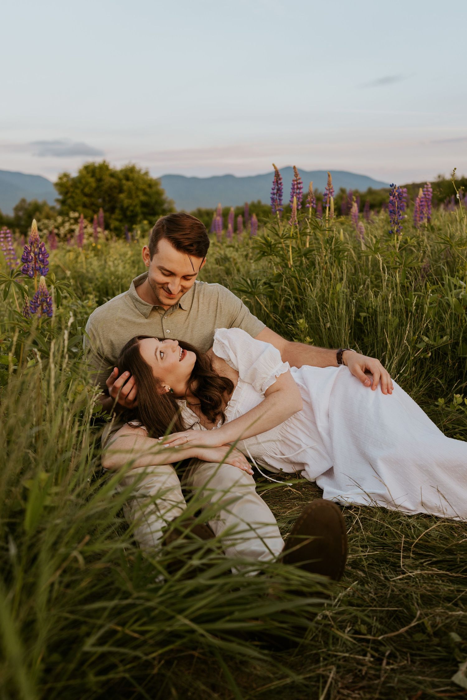 Couple relaxing in a lupine flower field at sunset, woman lying in man's lap smiling.