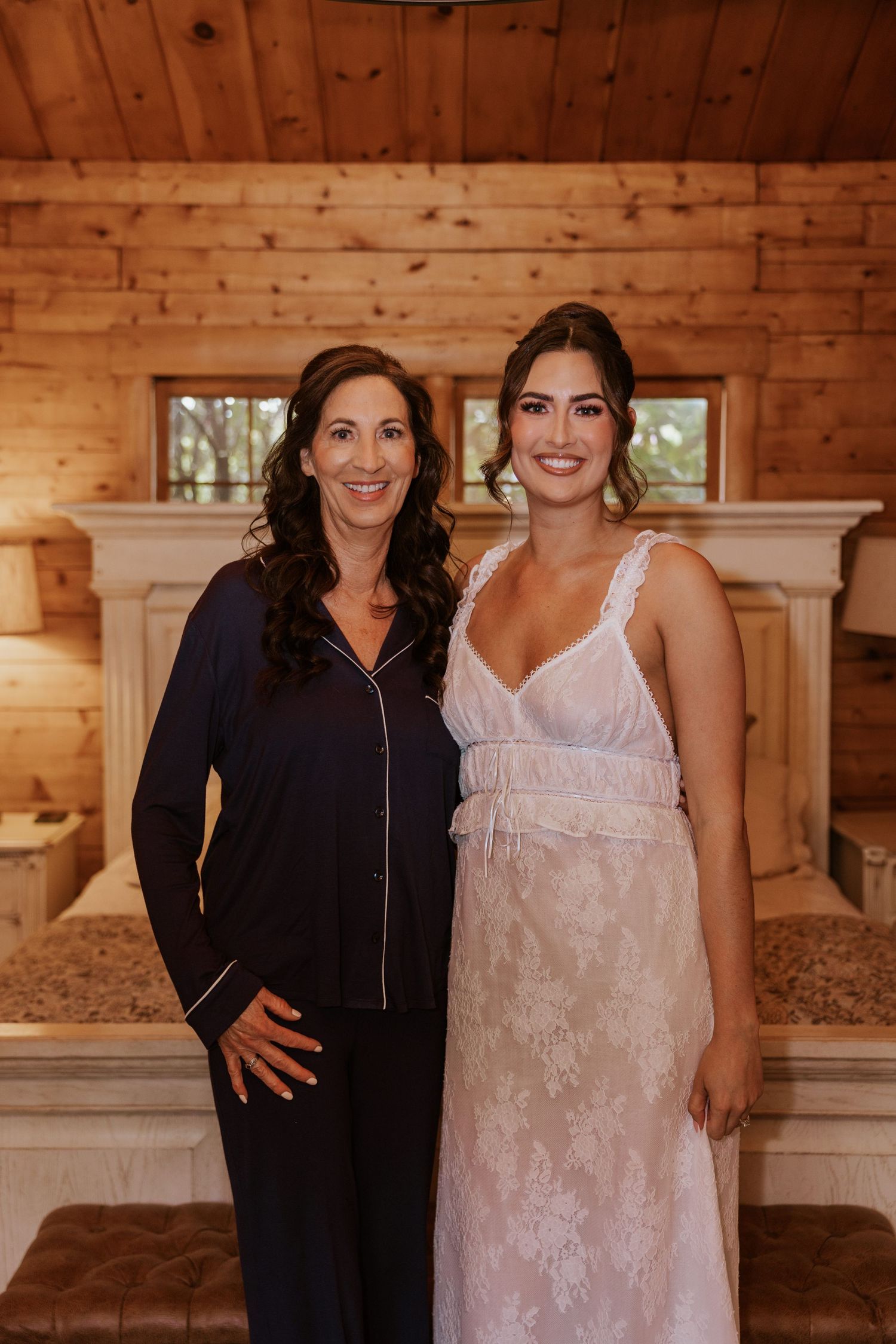 Two women smiling in a rustic log cabin, one in navy pajamas and one in a white lace bridal gown.