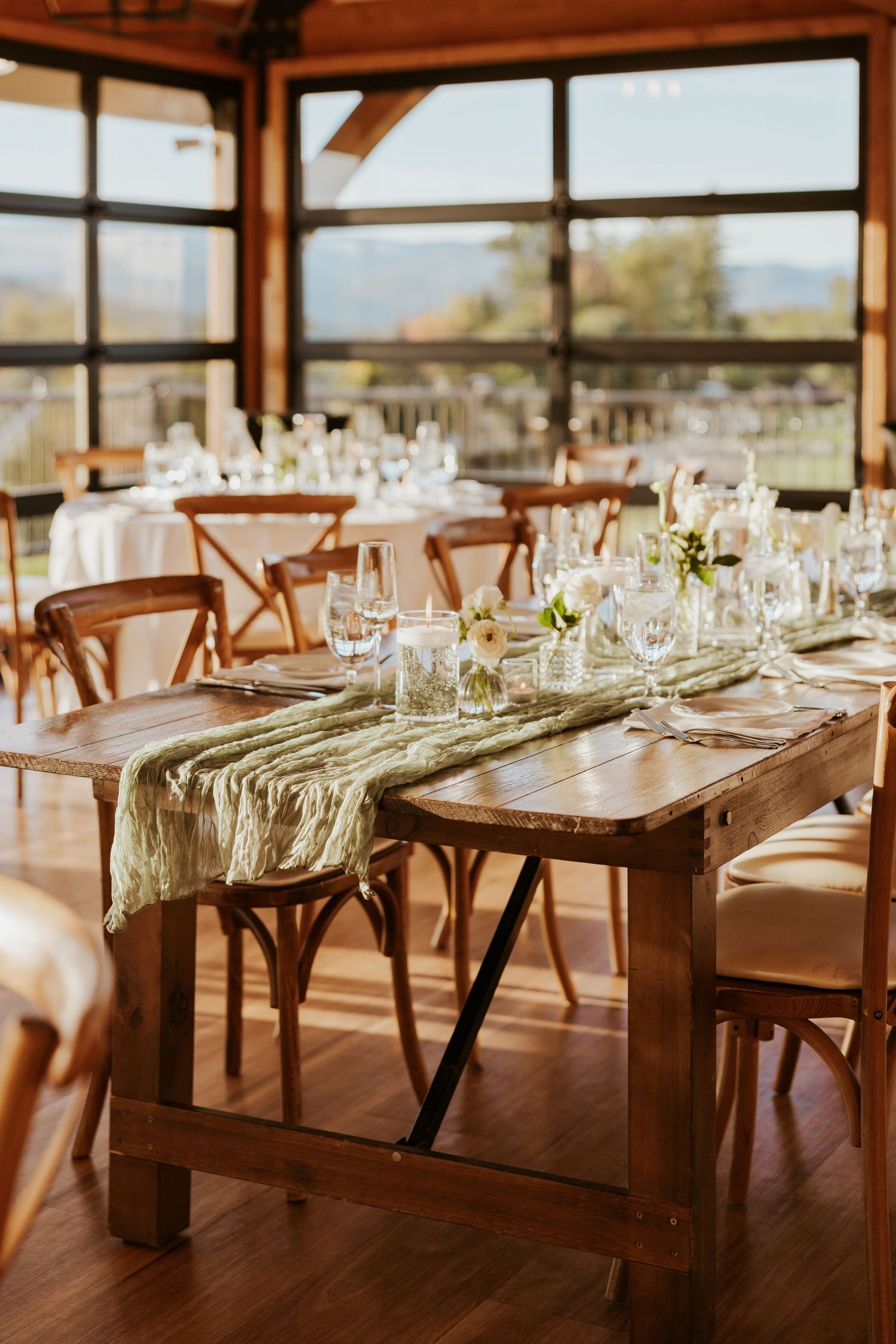 Rustic wooden farm tables with green velvet runners and floral centerpieces in sunlit wedding venue.