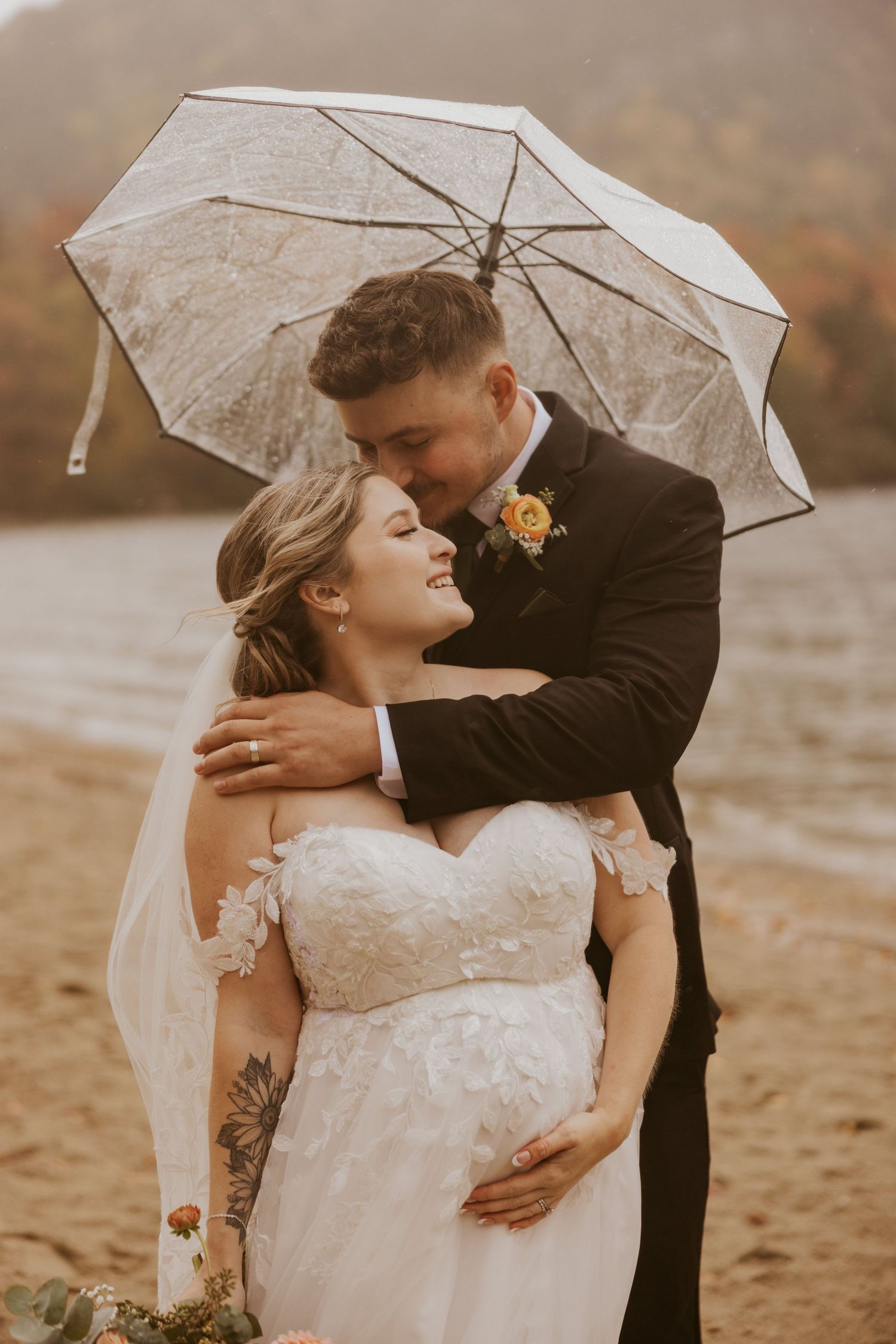 Groom tenderly embraces smiling bride under a lace umbrella on a misty fall lakeside wedding day.
