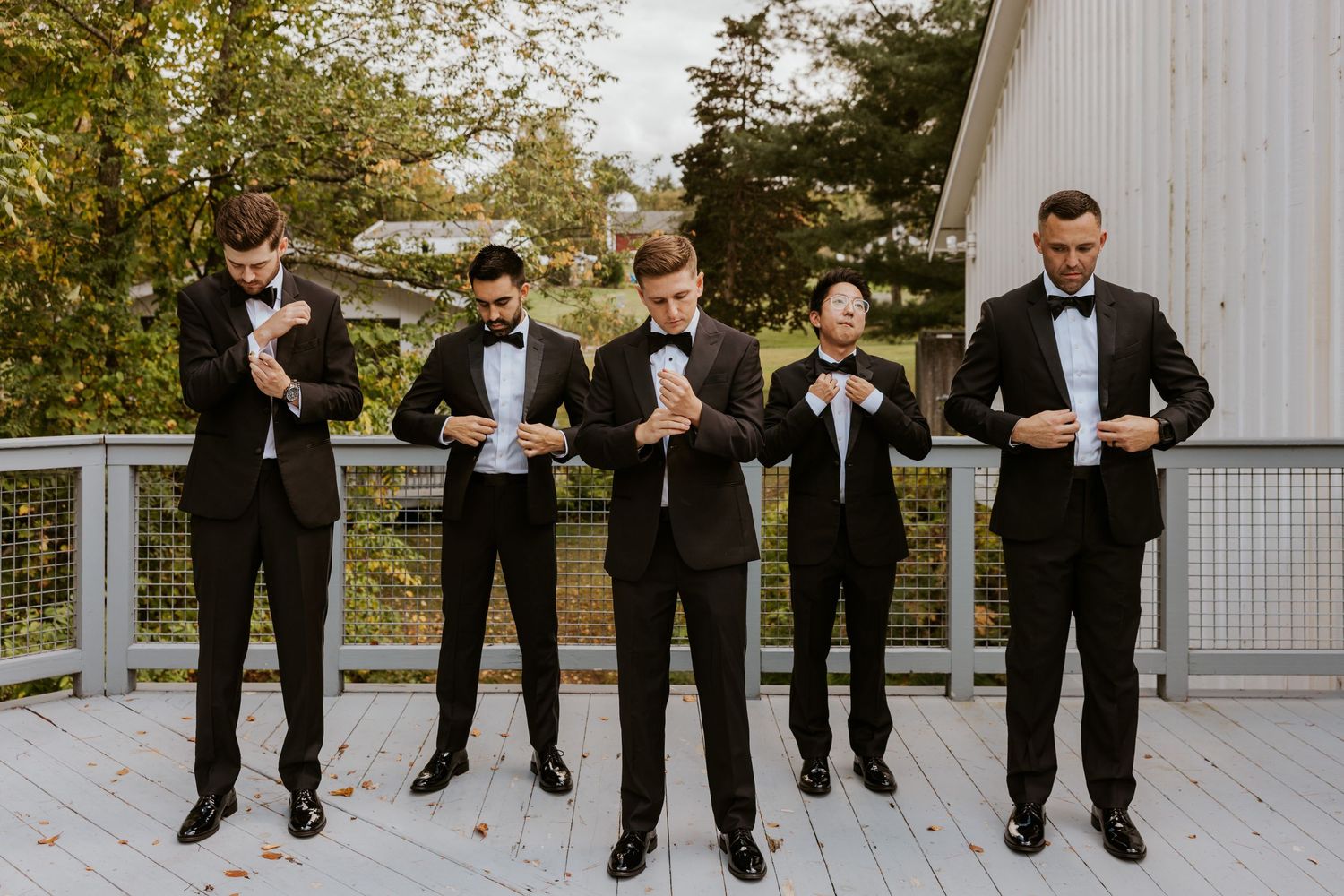 Five groomsmen in black tuxedos adjust their cufflinks together on an outdoor wooden deck.