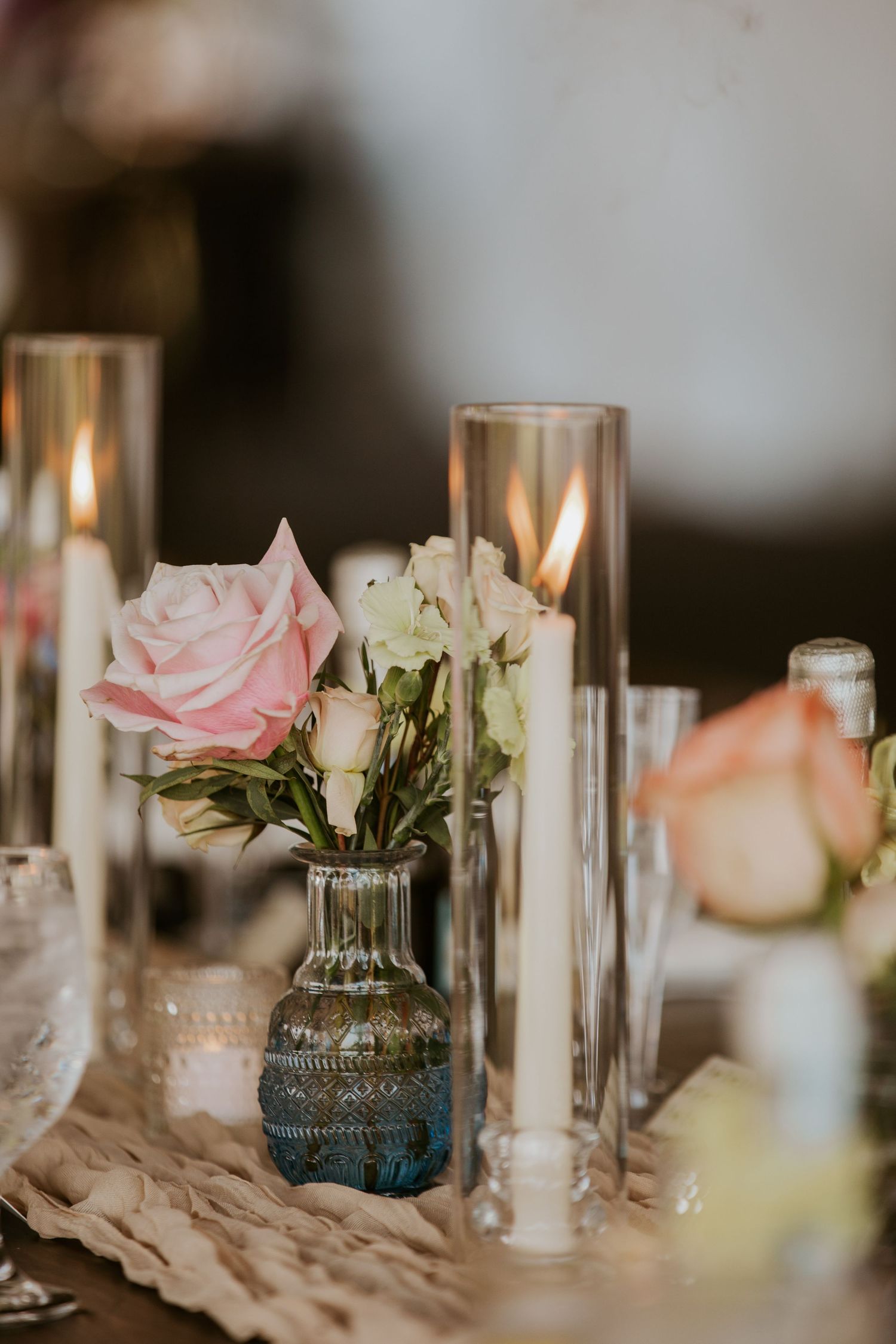 Elegant wedding table centerpiece with pink roses in blue glass bud vase surrounded by tall glass candle holders.