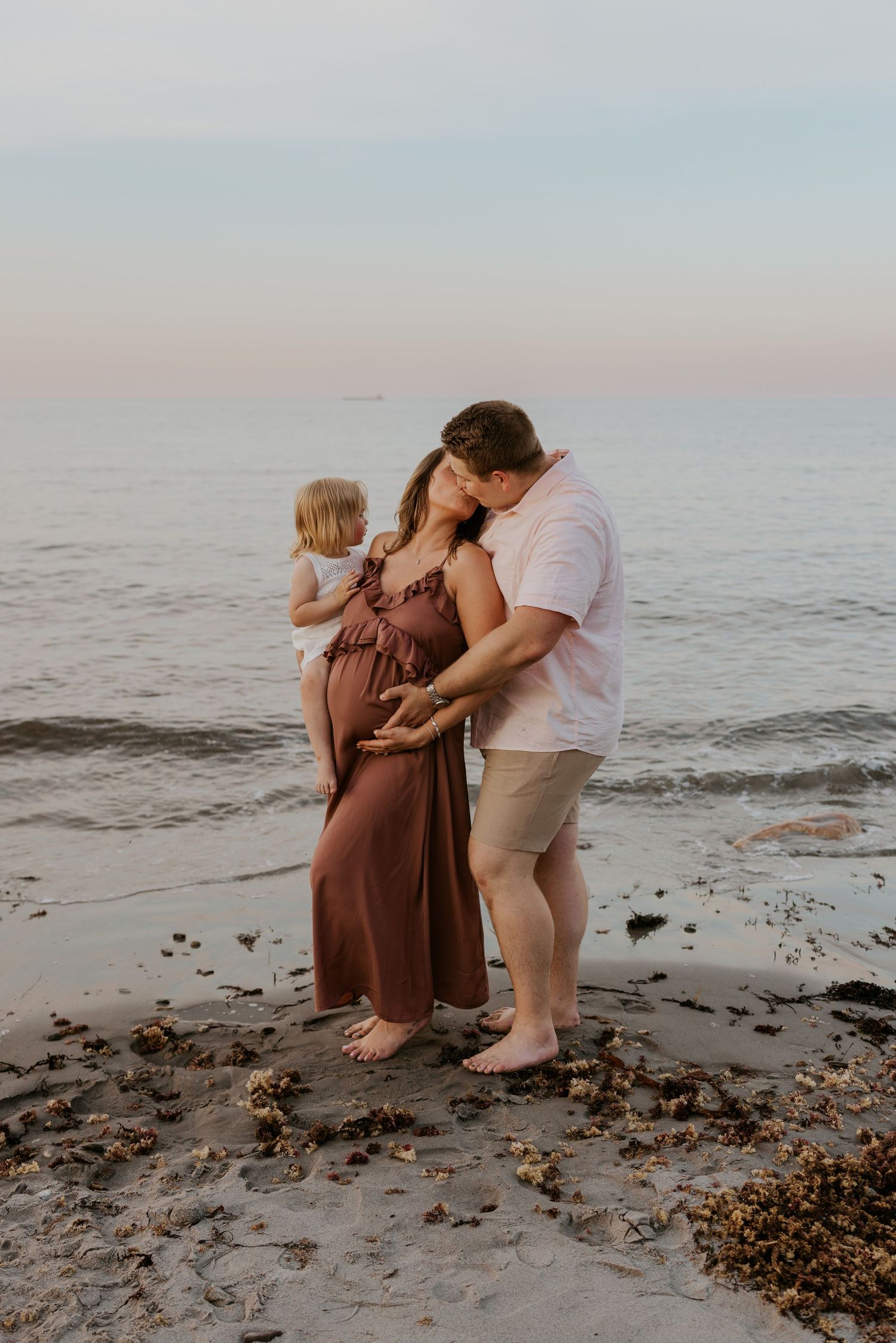 Couple tenderly embracing pregnant belly on beach at dusk with toddler standing nearby.
