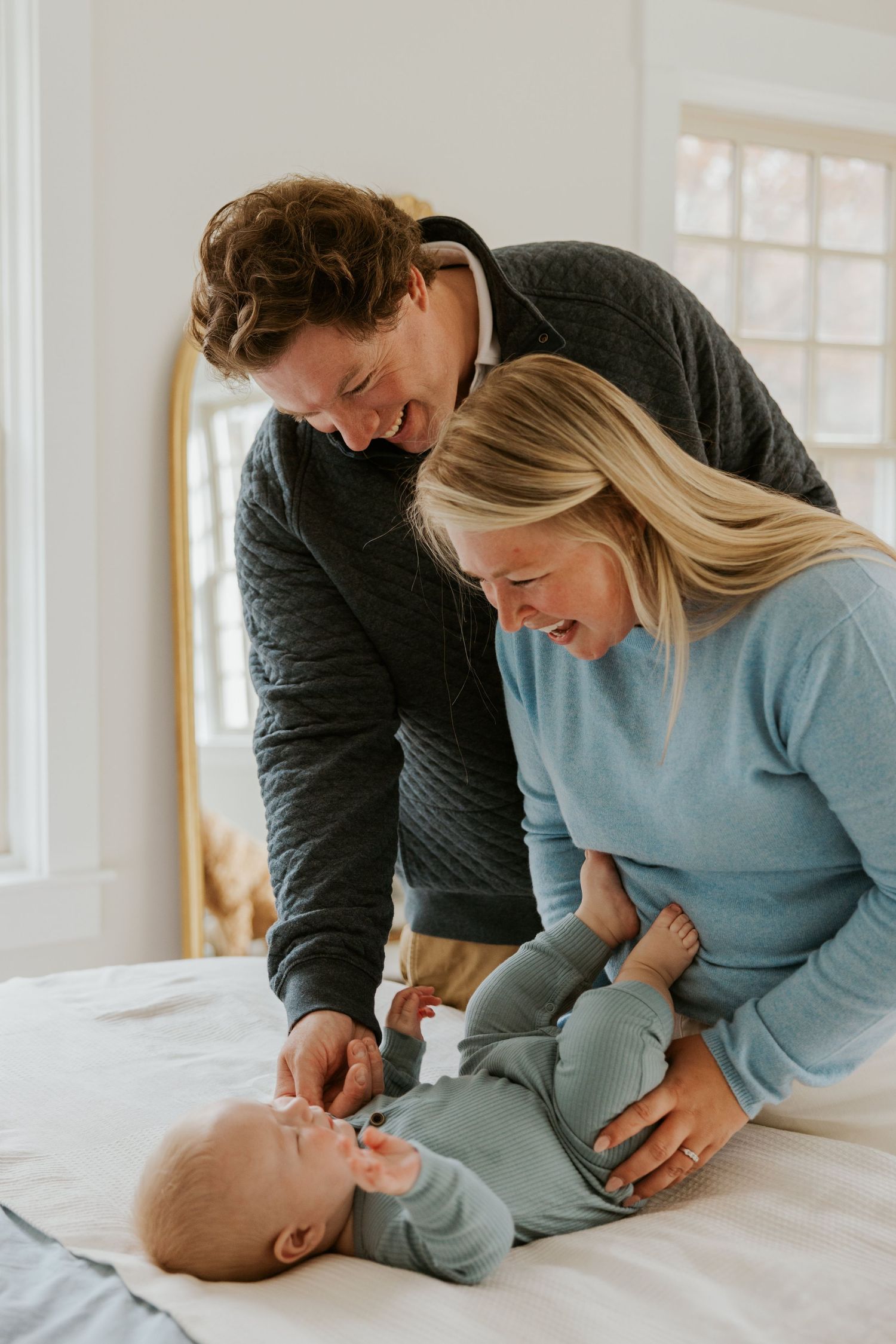Laughing parents in coordinating blue and gray outfits playing with baby on white bed.