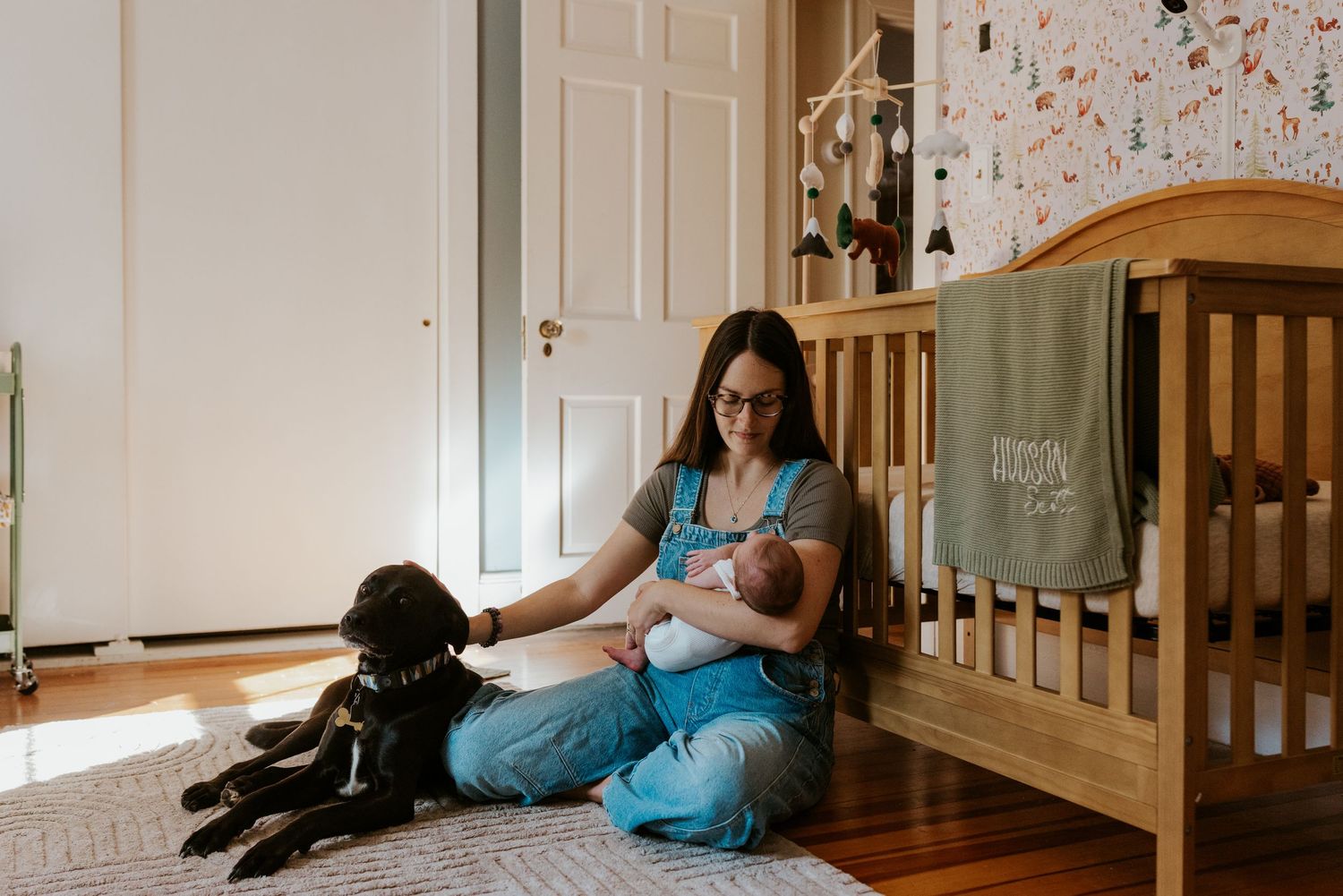Mother sitting on floor nursing newborn baby beside wooden crib with black dog nearby in nursery.