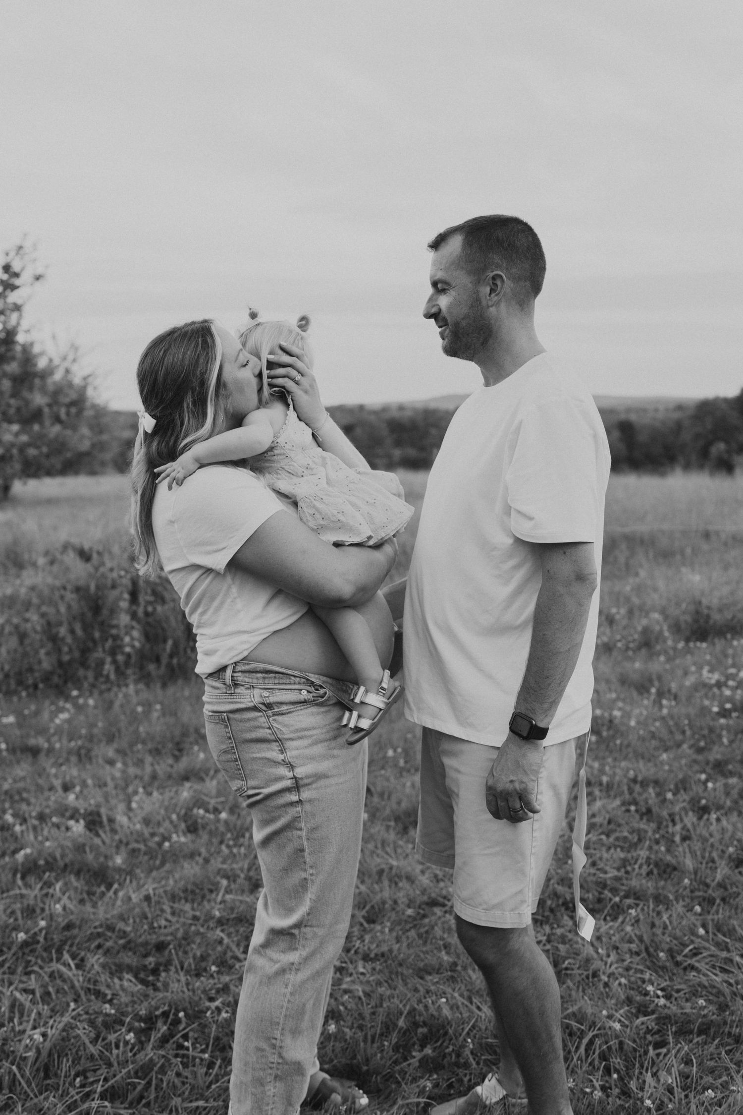 Black and white photo of a pregnant woman kissing her toddler while her smiling husband stands beside them outdoors.