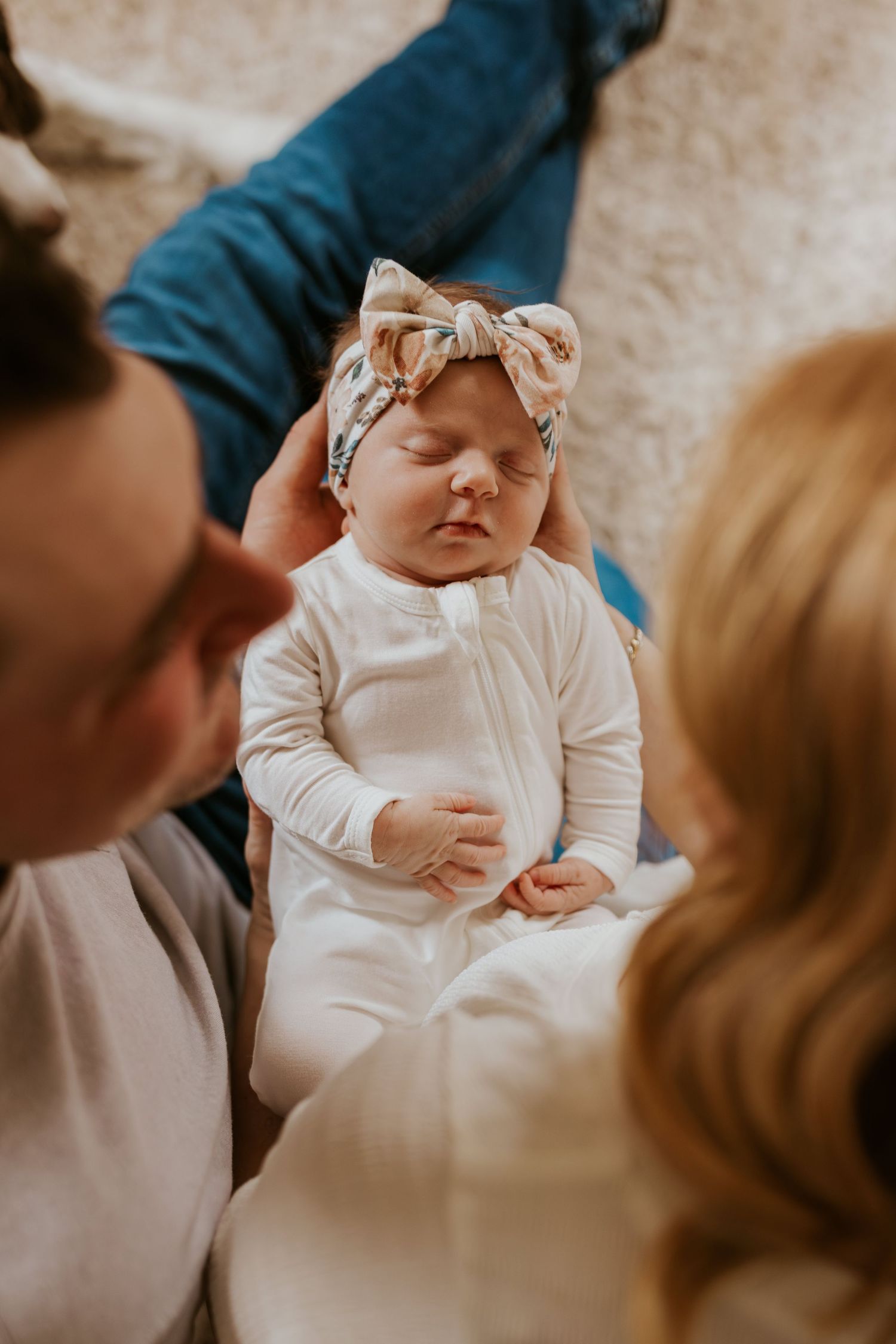 Overhead view of parents gazing down at their sleeping newborn baby girl wearing a floral bow headband.