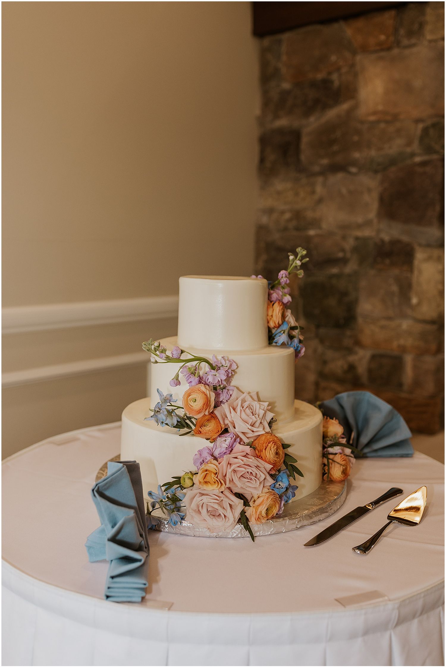 Elegant white wedding cake decorated with fresh pink roses and blue accents on white tablecloth.