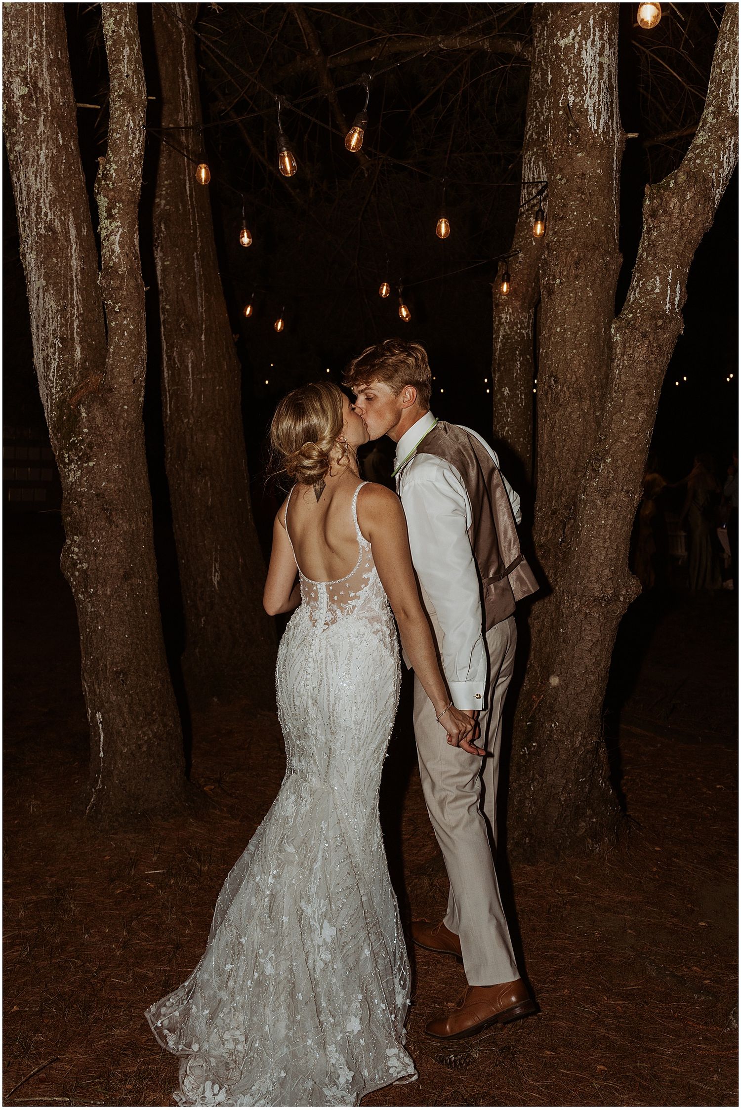 Couple sharing an intimate moment among trees with hanging lights at night.