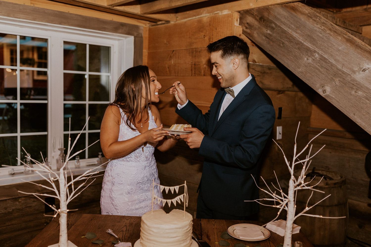Couple sharing laughter while cutting their wedding cake at a rustic reception.