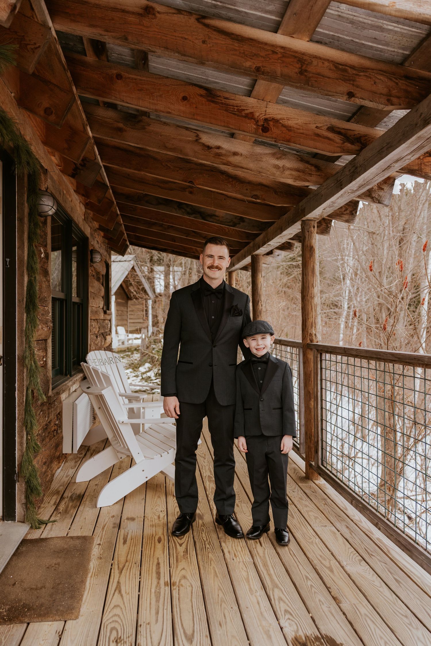 Groom and young ring bearer in matching black suits posing on a snowy rustic wooden porch together.