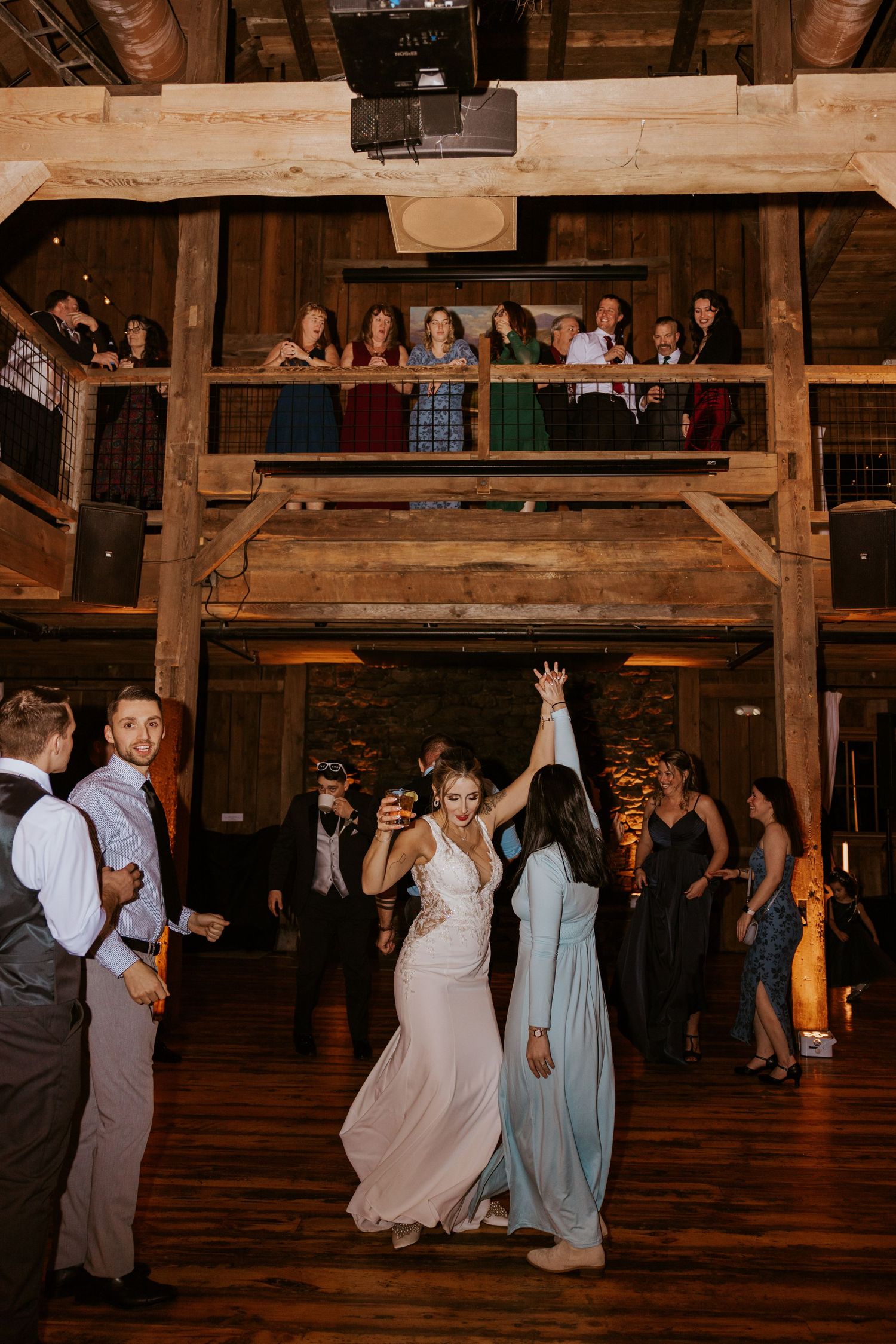 Bride dancing joyfully with guests in a rustic barn wedding venue with wooden beams and balcony onlookers.