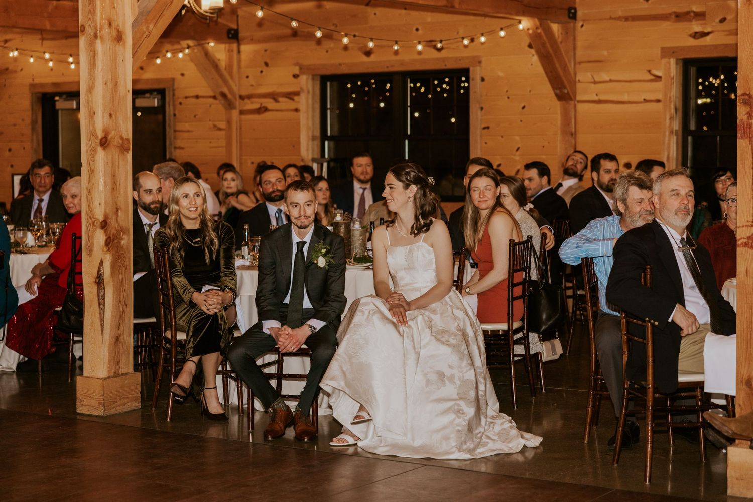 Wedding guests dance and celebrate on a rustic wooden dance floor illuminated by warm string lights overhead.