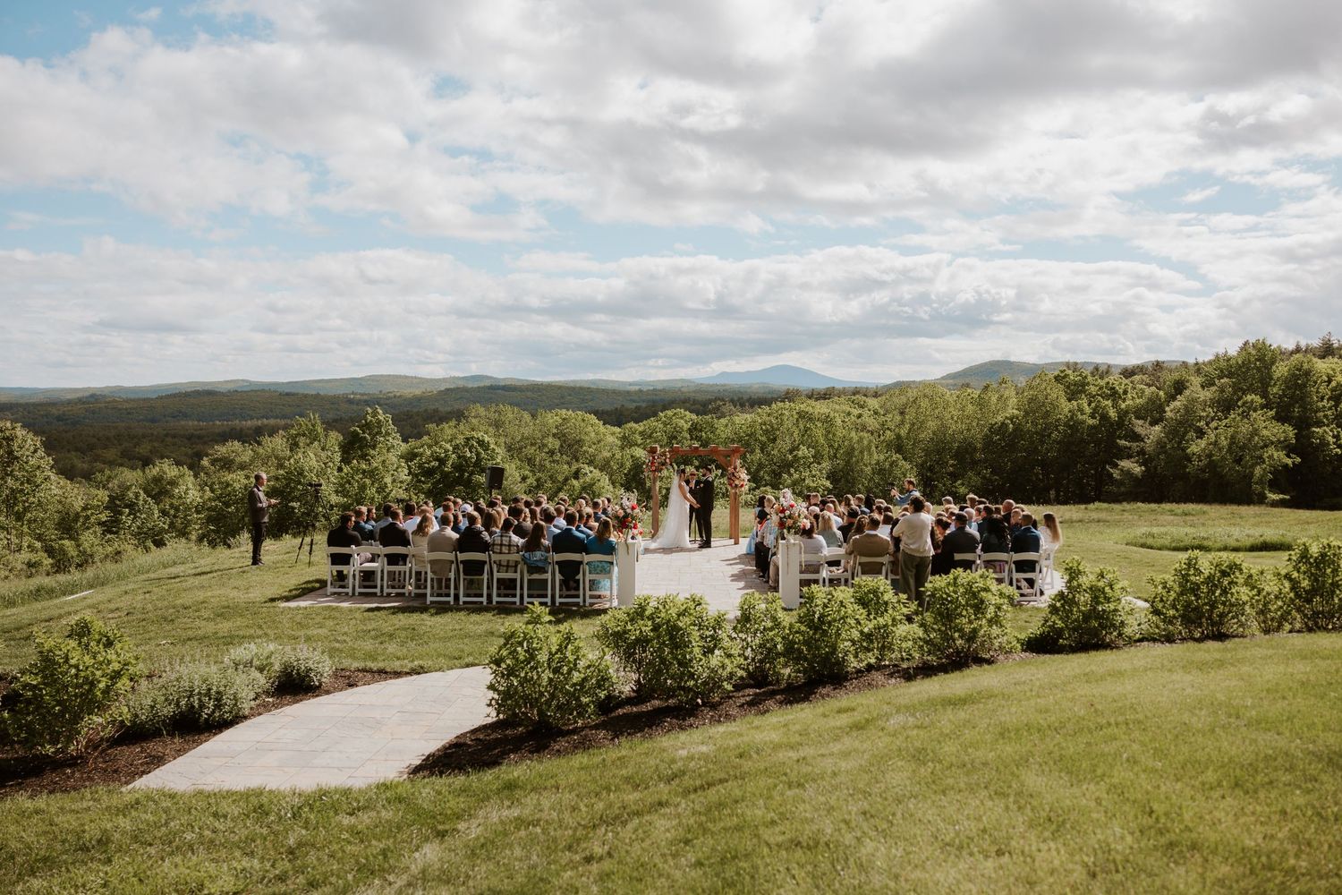 Outdoor wedding ceremony on a lush green hillside with mountain views and guests seated in rows under a cloudy sky.