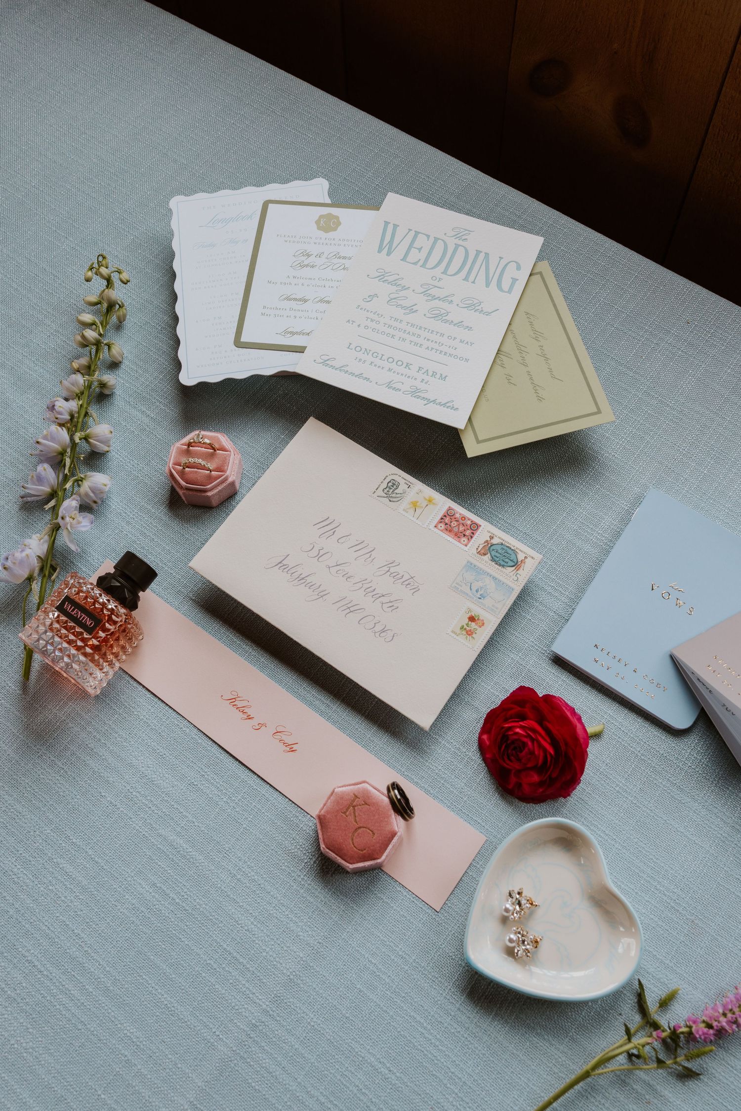 Elegant wedding invitation suite with floral accents, perfume, red rose, earrings, and wax seal on a soft blue surface.