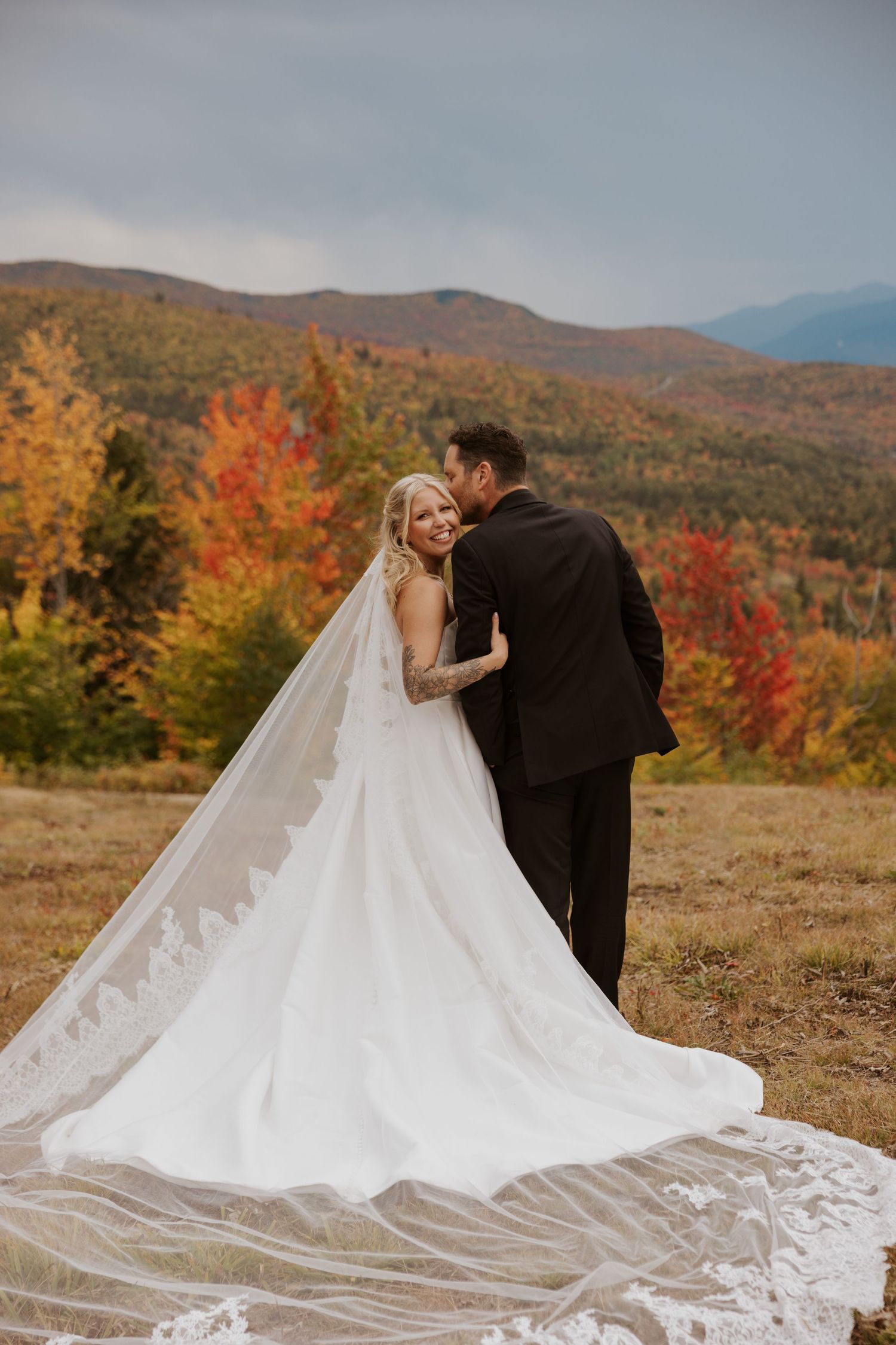 Bride and groom embrace in autumn mountain landscape, her lace cathedral veil flowing dramatically in the breeze.