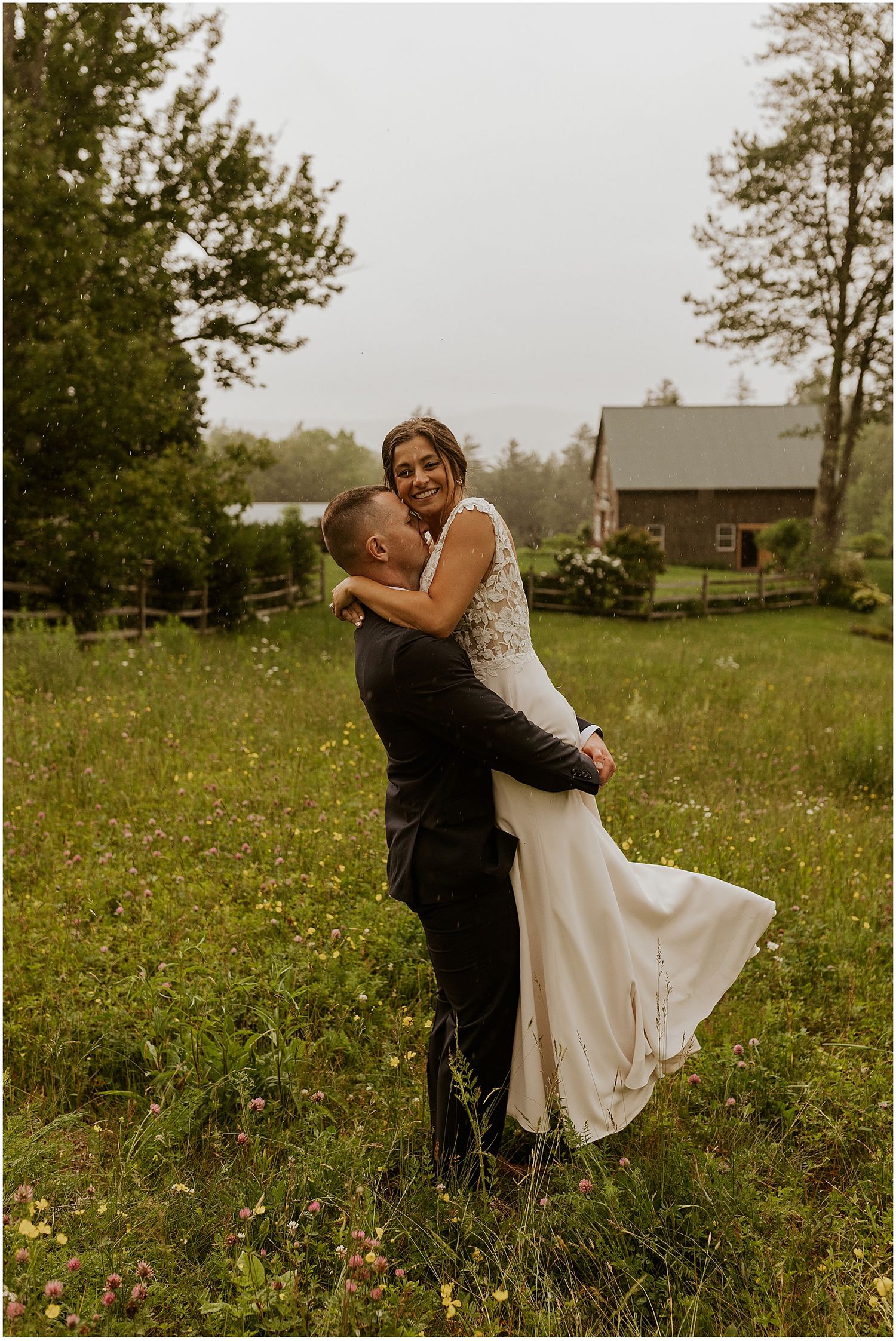 A couple spins joyfully in a field of wildflowers near a countryside barn.