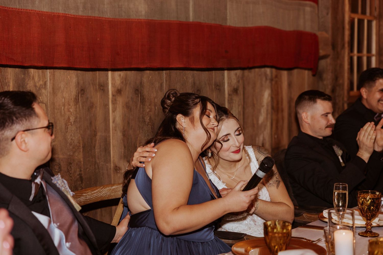 Two bridesmaids share an emotional hug during wedding reception speeches at a rustic barn venue.