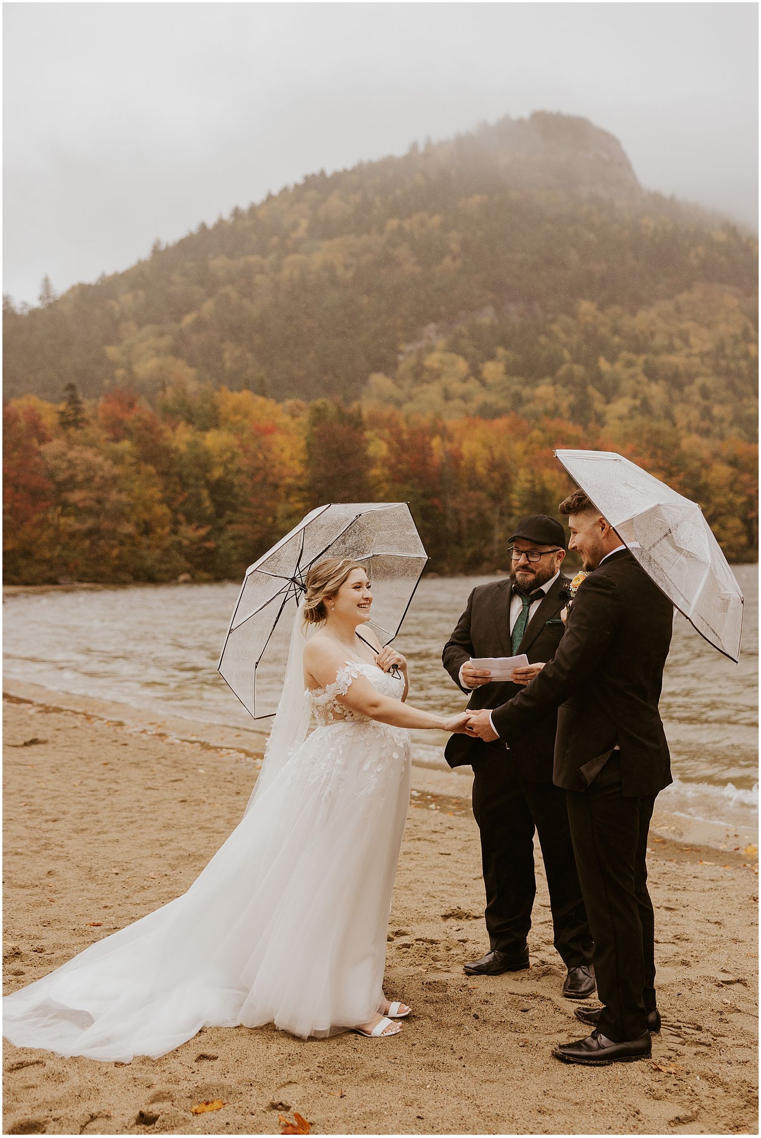 Wedding ceremony on a beach with clear umbrellas and misty mountain scenery.