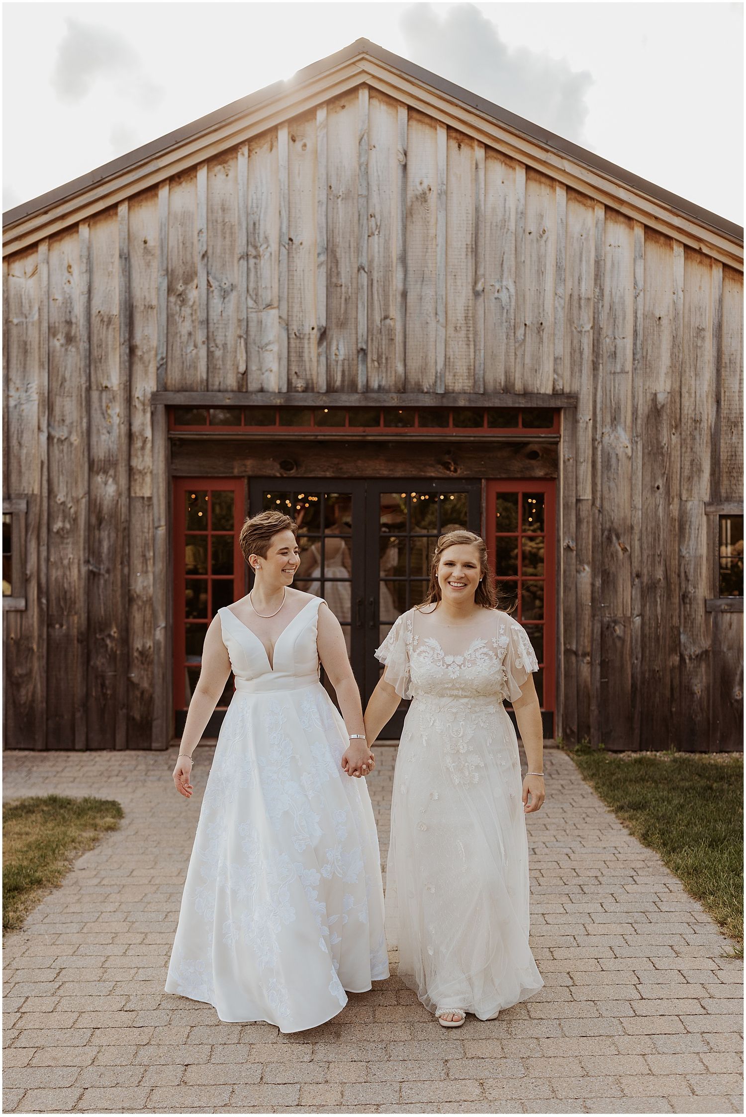 Two people in elegant white wedding dresses hold hands while standing in front of a rustic wooden barn.