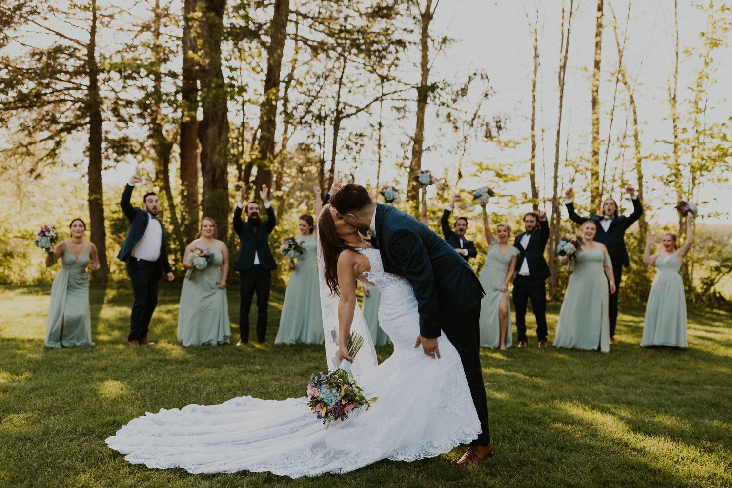 The groom is dipping the bride and kissing her as the wedding party cheers and jumps behind them.